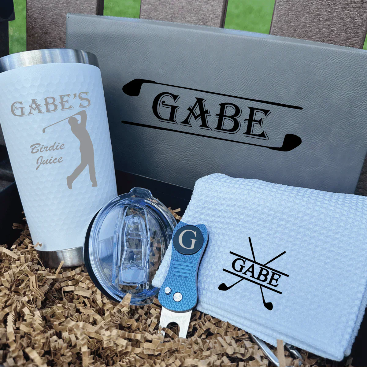 Assortment of personalized golf gifts under $100 including an engraved decanter, custom golf towel, monogrammed shoe bag and a magnetic cart koozie arranged on turf