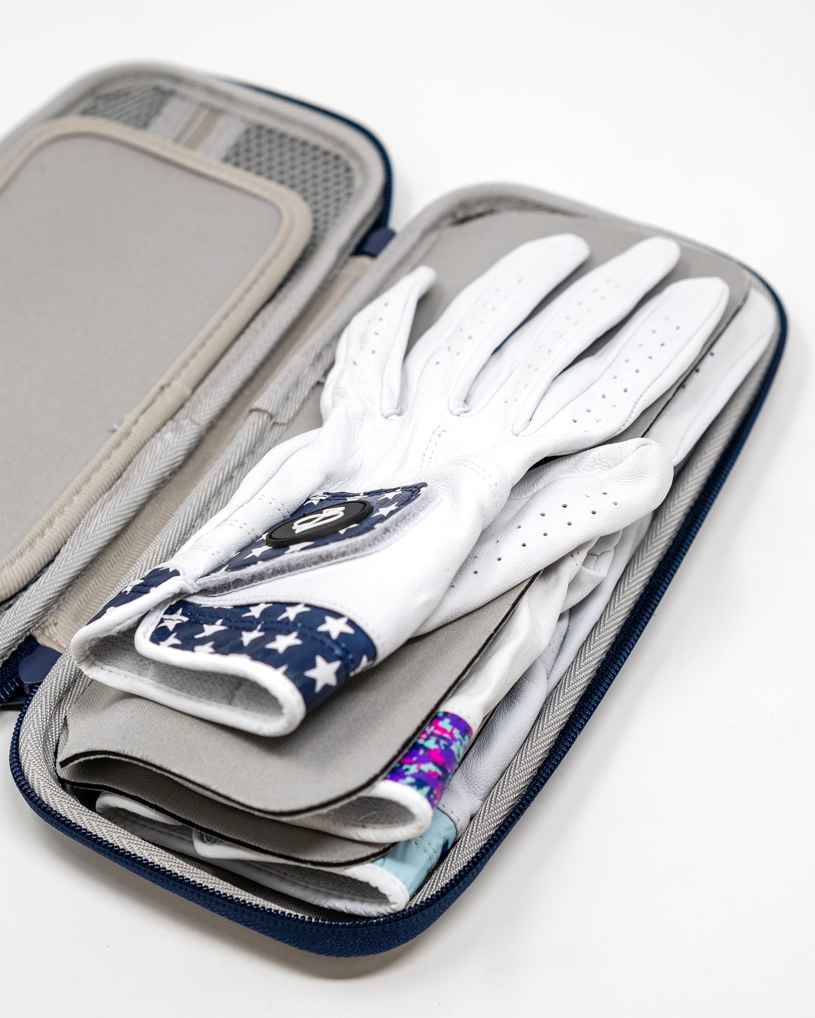 White leather golf glove with navy star pattern and colorful wrapped grip, displayed inside navy and gray glove caddy with organized compartments.