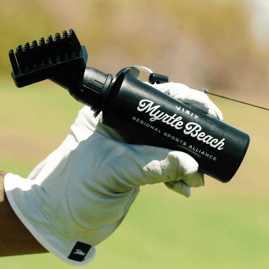 Black golf club brush with engraved "Myrtle Beach" and "Regional Sports Alliance" text, attached to golf bag with retractor clip, held in gloved hand over green course.