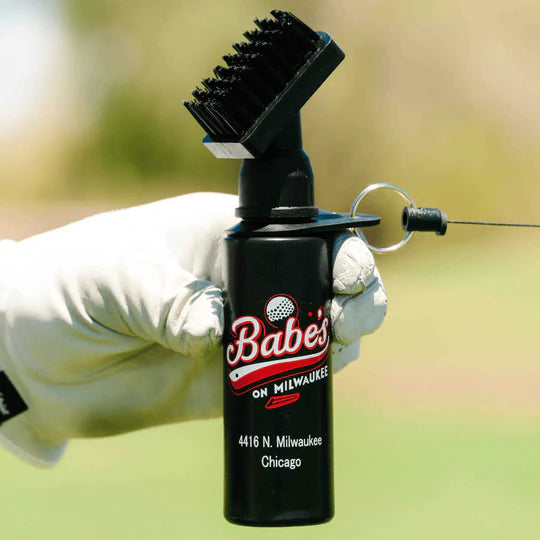 Black golf club brush with nylon bristles and attached water spray bottle, held in hand with golf ball, showing retractor clip and logo placement on bottle body