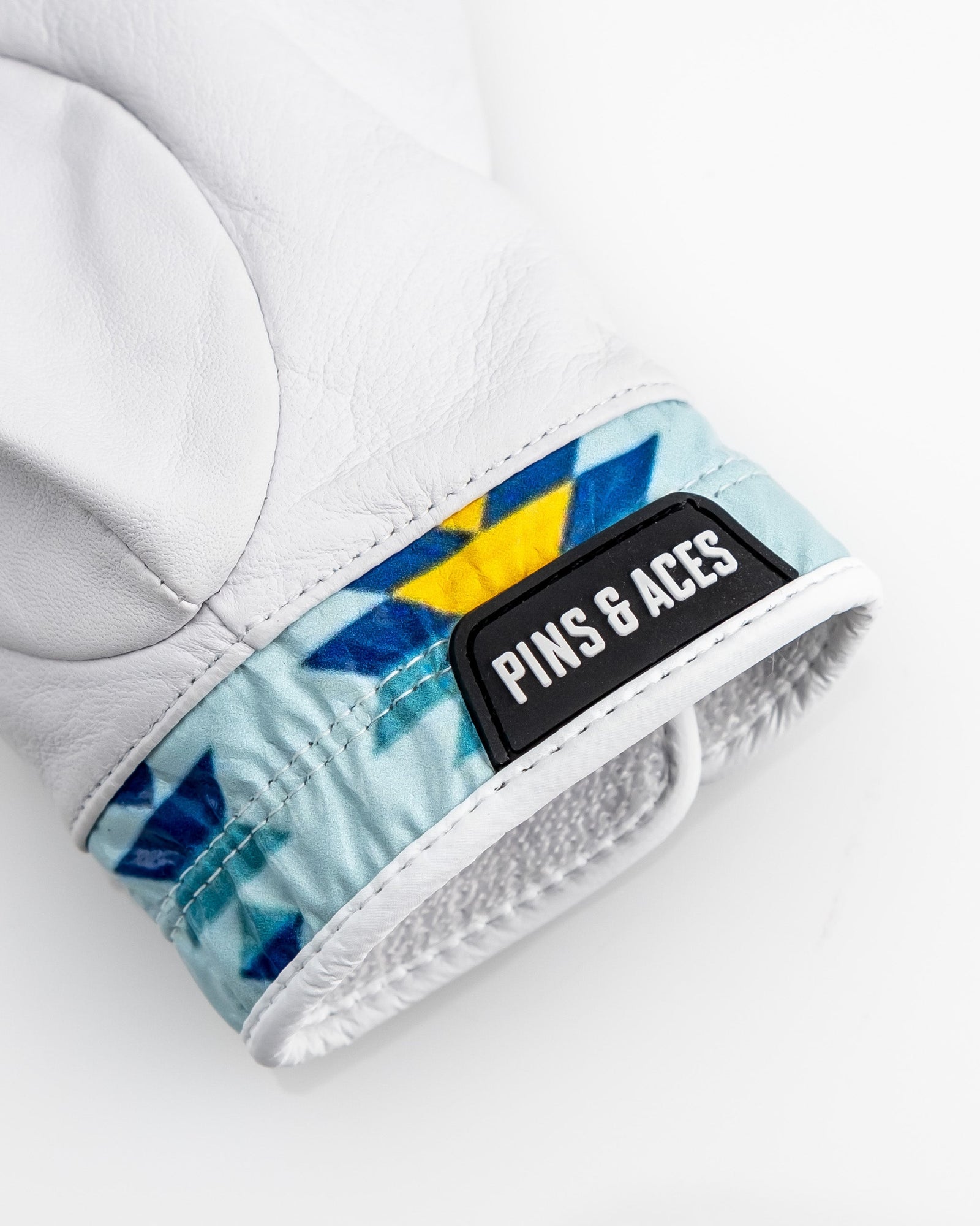 White leather golf glove with blue and yellow geometric pattern on cuff, displaying black Pins & Aces brand patch and magnetic ball marker detail.