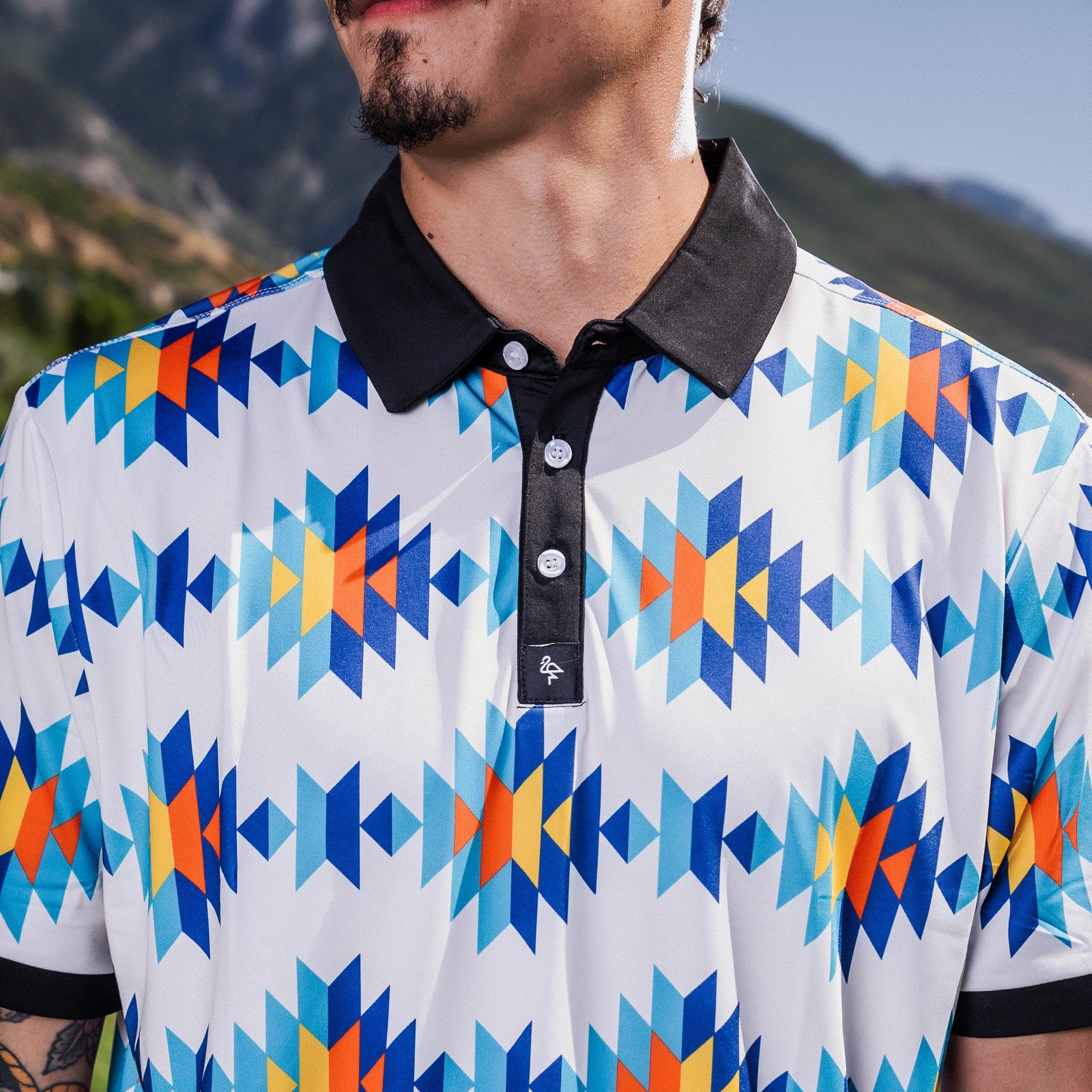 White Aztec polo with teal, orange & navy diamond pattern, 4‑way stretch, UPF 50+, moisture‑wicking, anti‑odor & wrinkle resistance.