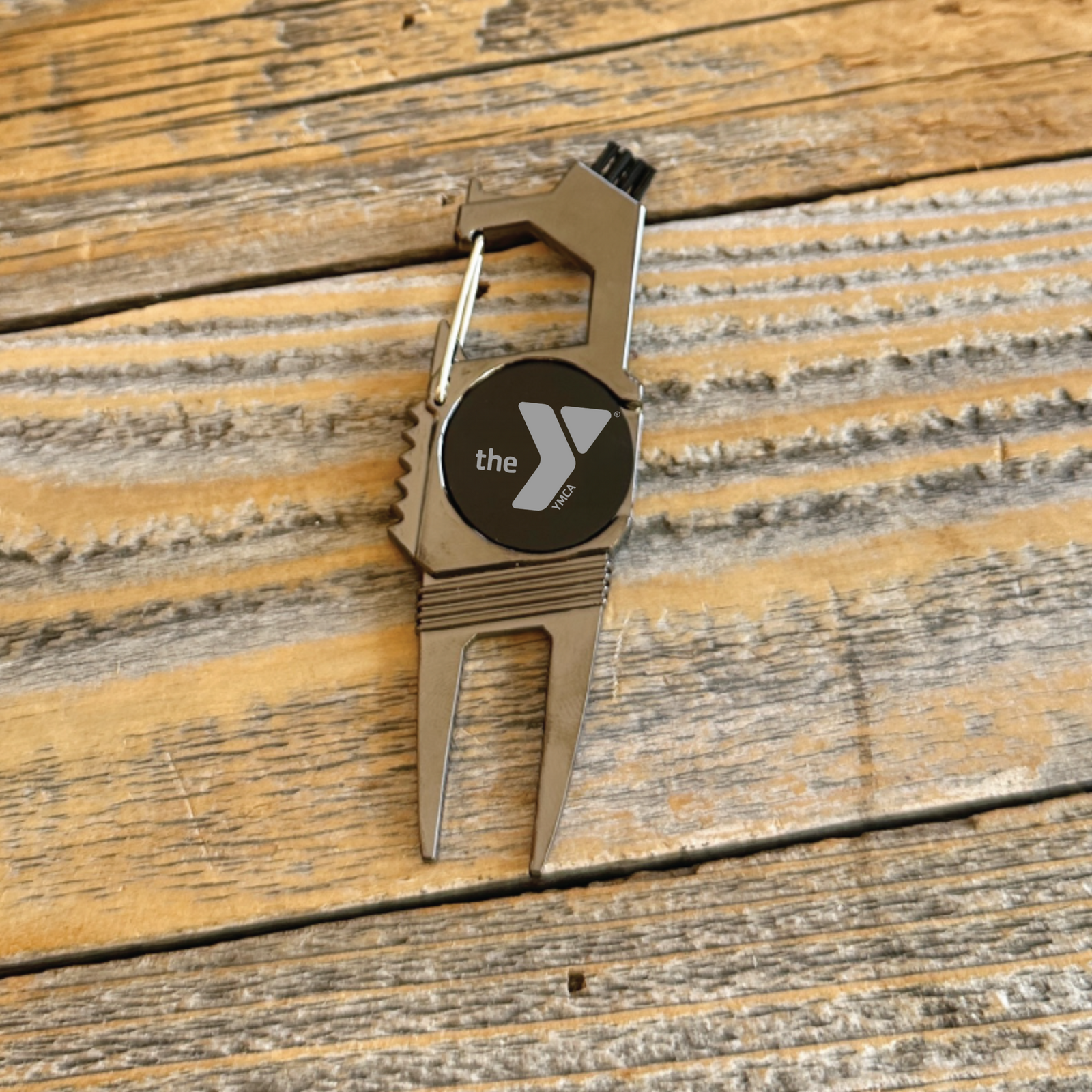 Tan divot tool with black circular face displaying "the Y" logo and brush bristles at top, displayed on weathered wood surface