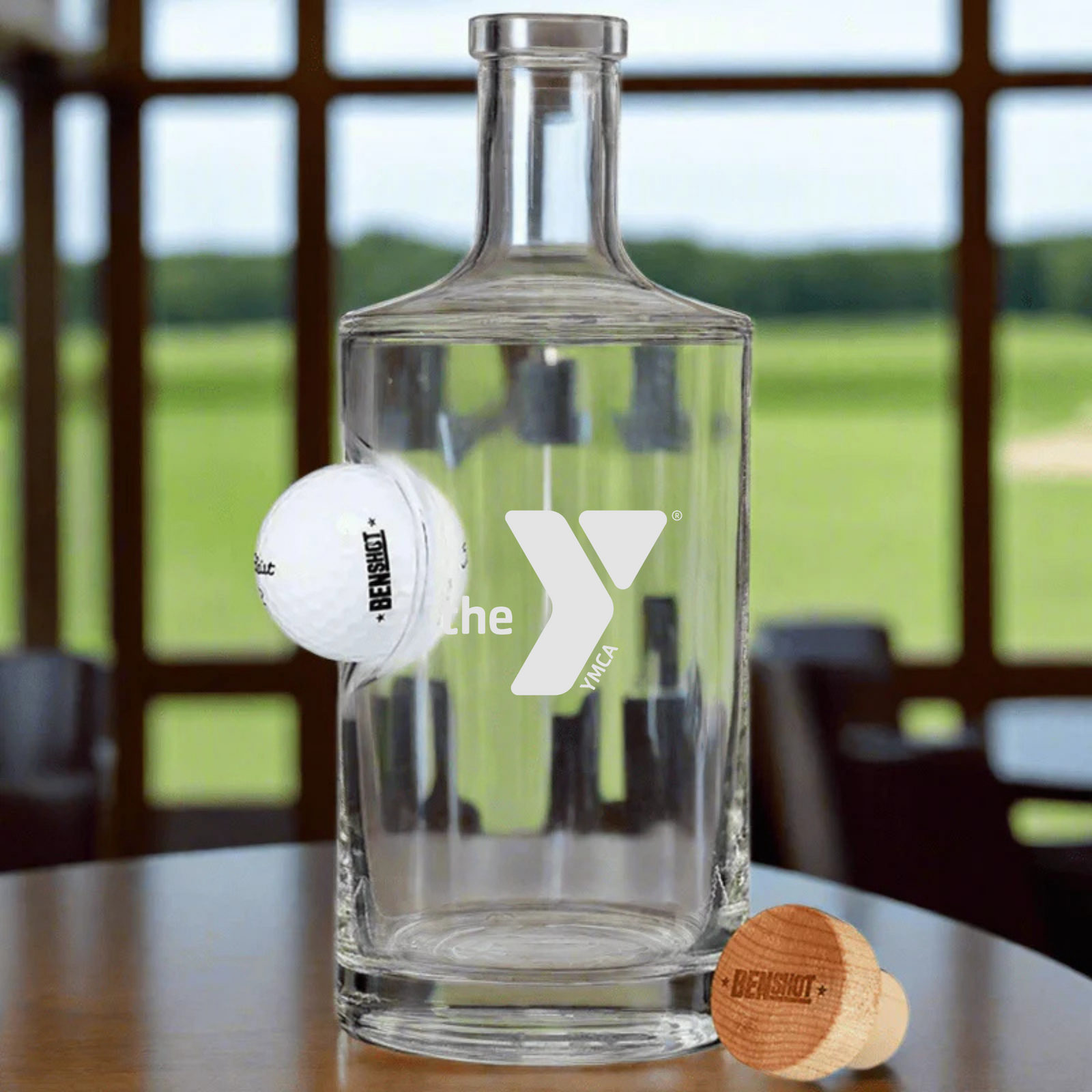 Clear glass decanter with white logo and wooden stopper engraved with sample text, displayed on table with golf ball and clubhouse view in background