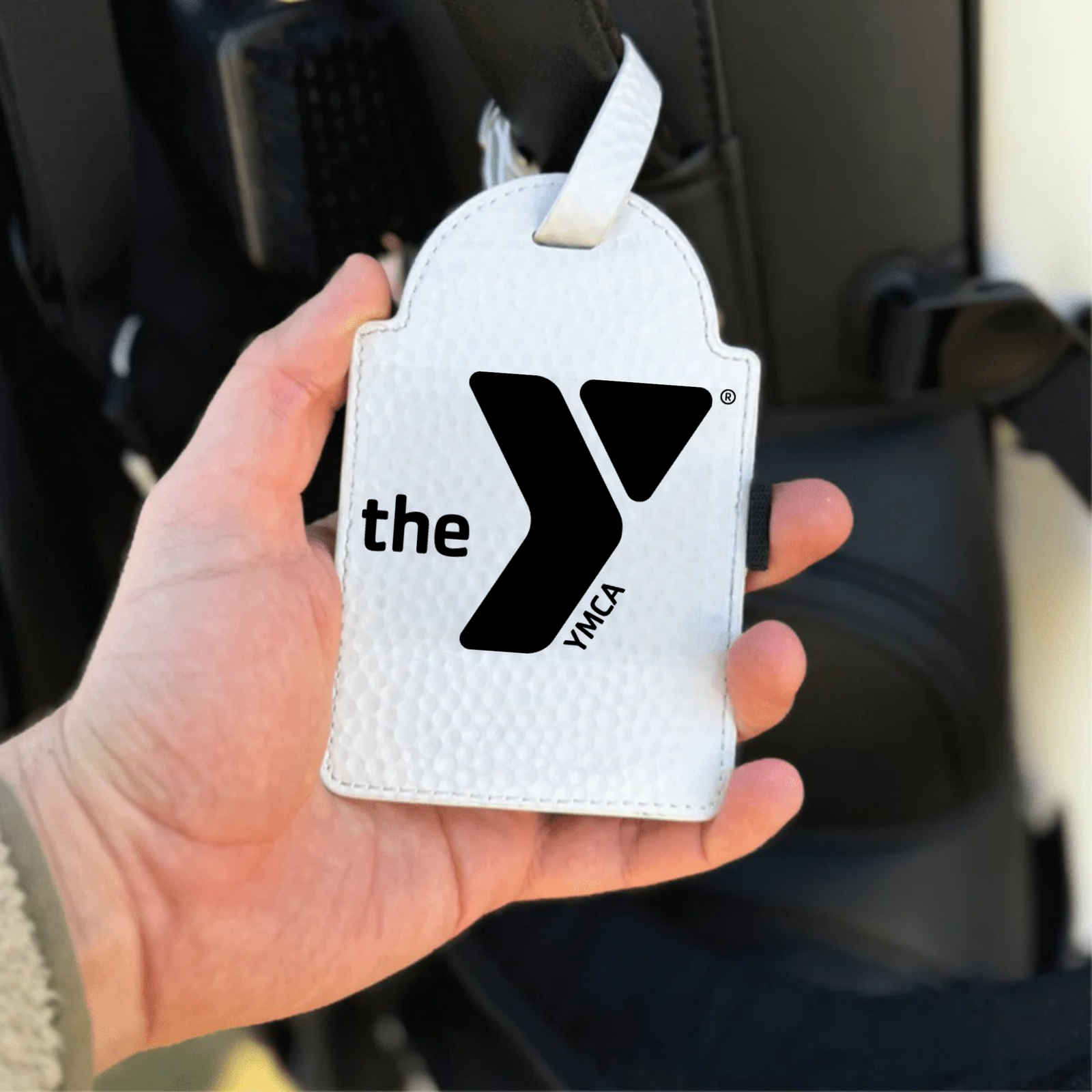 White golf bag tag with black YMCA logo held in hand, featuring attached white strap for attachment to golf bag handle