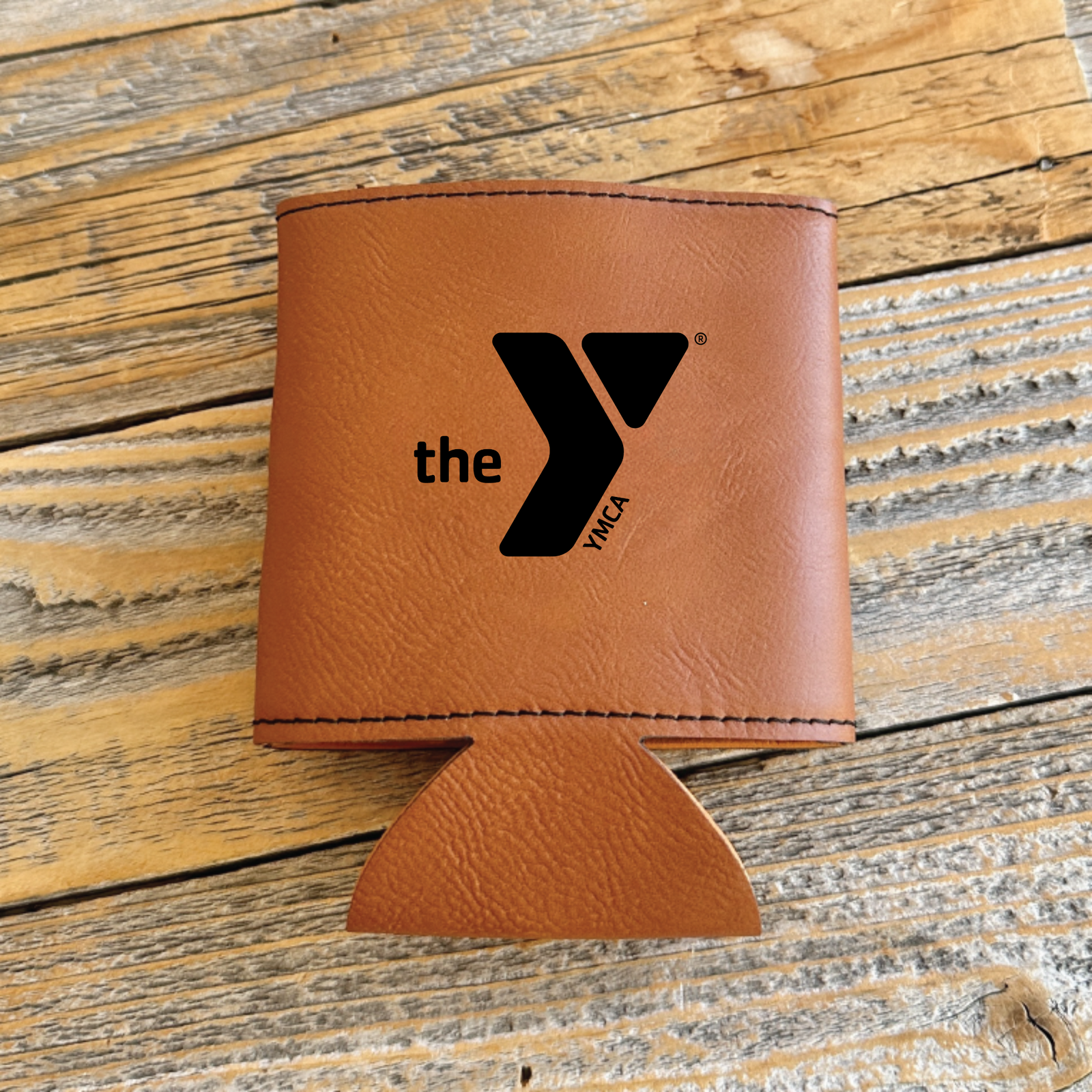 Tan leather koozie with embroidered YMCA logo and "the" text, displayed flat on rustic wood surface