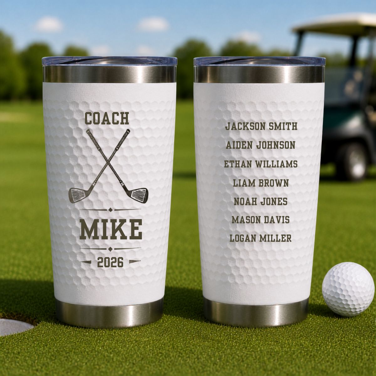 White dimpled golf ball tumbler for a golf coach, front engraved COACH MIKE 2026 with crossed clubs, back engraved with seven player names, on a putting green
