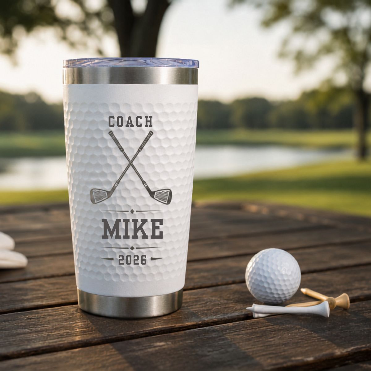 Front of a white golf coach tumbler engraved COACH, crossed clubs, MIKE and 2026, resting on a wooden table beside a golf ball and tee