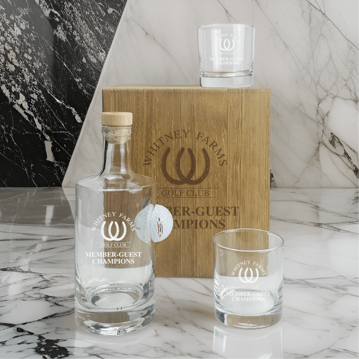 Wood box with laser-engraved Whitney Farms Golf Club Member-Guest Champions logo, paired with decanter, stopper, and two matching glasses on marble surface