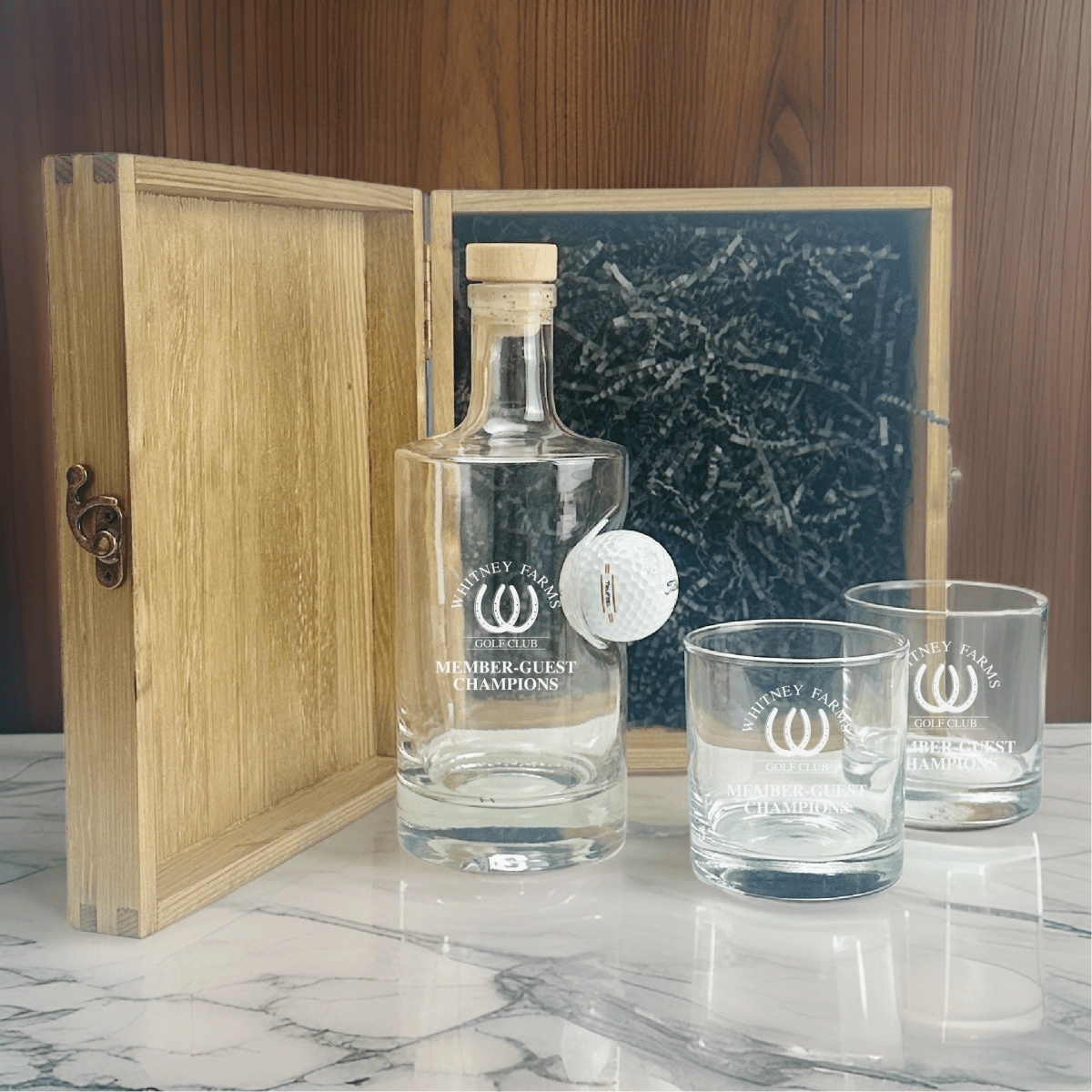 Golf decanter gift set displayed with open wooden box, showing engraved decanter with embedded golf ball, two matching glasses etched with logo, and decorative ball marker