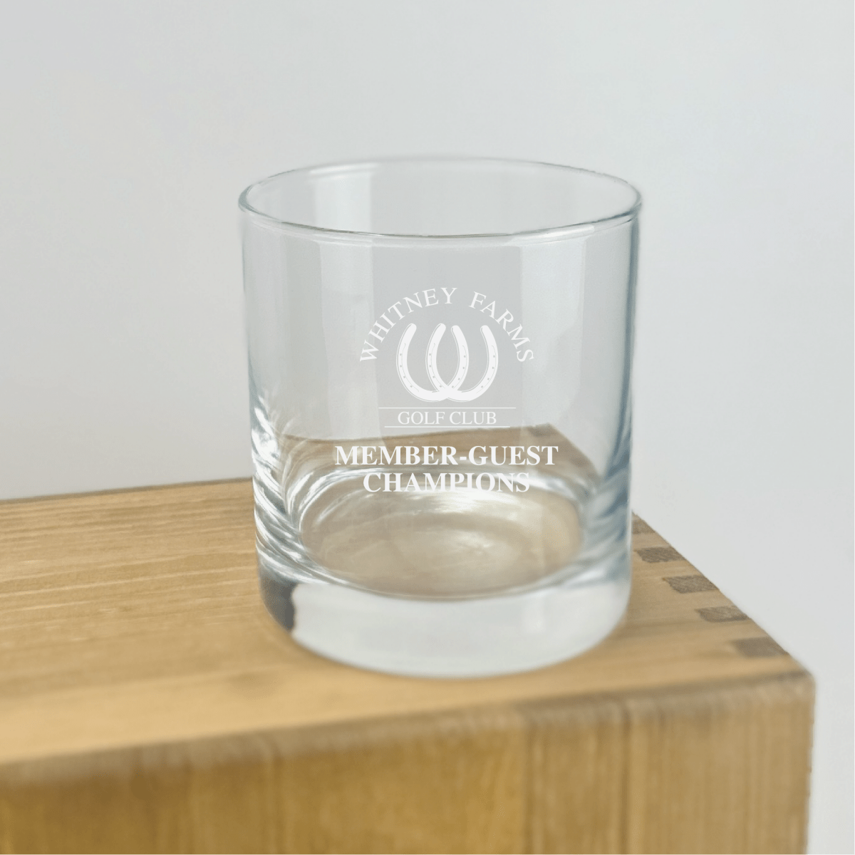 Etched rocks glass displaying Whitney Farms Golf Club logo and "Member-Guest Champions" text, sitting on wooden base with sample engraving