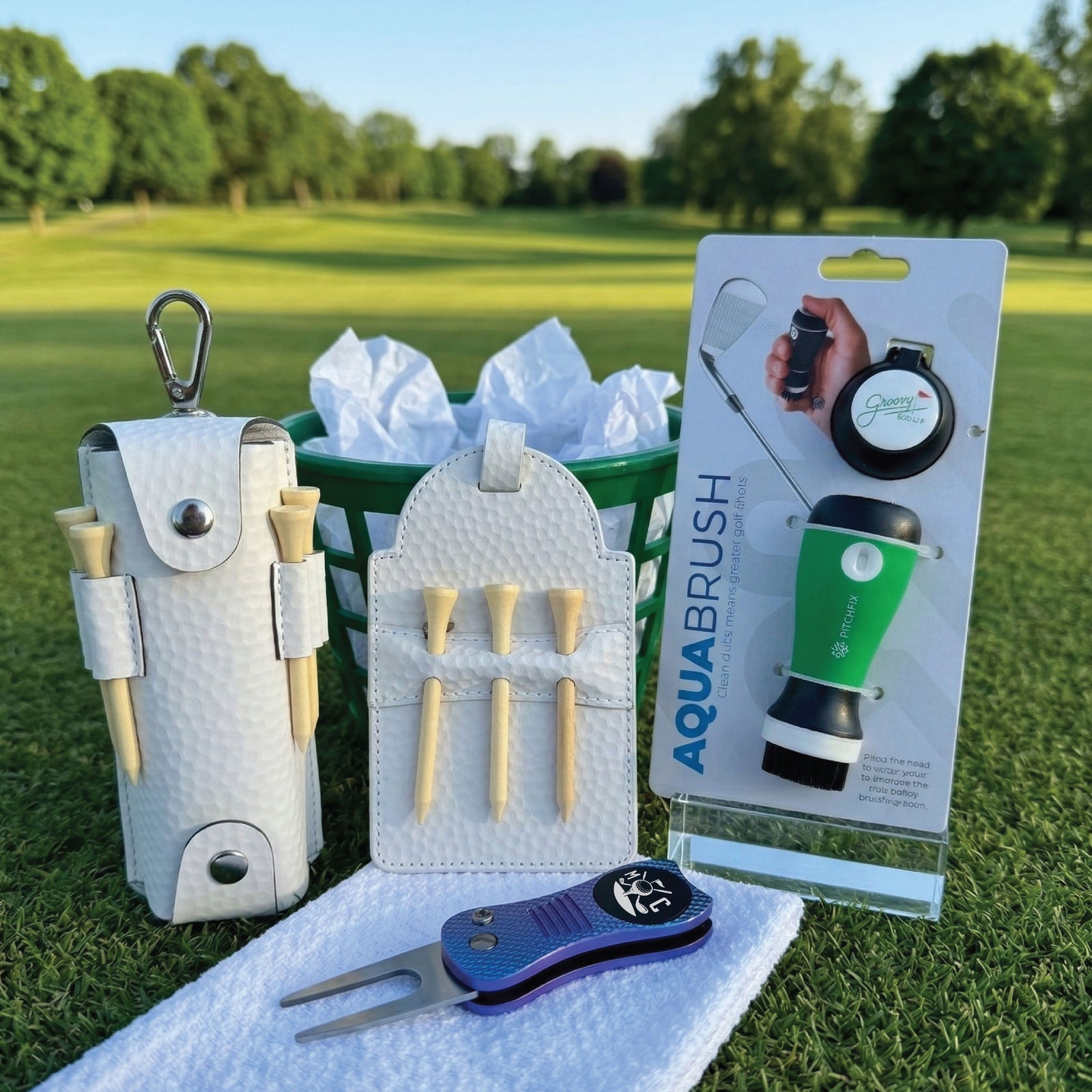 Golf gift set displayed on course grass including white tee pouch, peg holder, ball marker divot tool, towel, brush, tumbler, and packaging on green background.