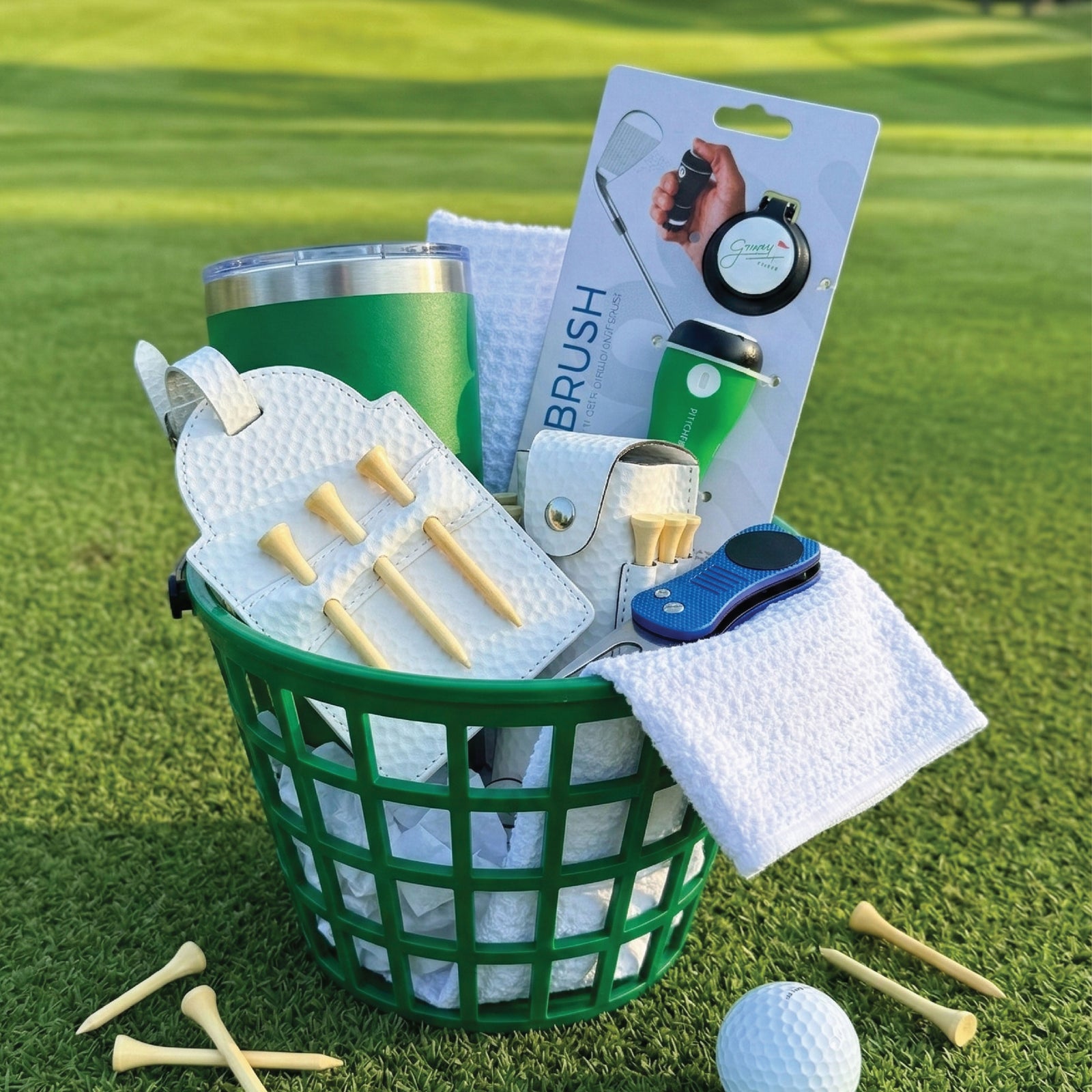 Golf gift basket with green mesh container holding a green insulated tumbler, white waffle towel, wooden tees, ball marker, divot tool, brush, and blue ball pouch on grass background