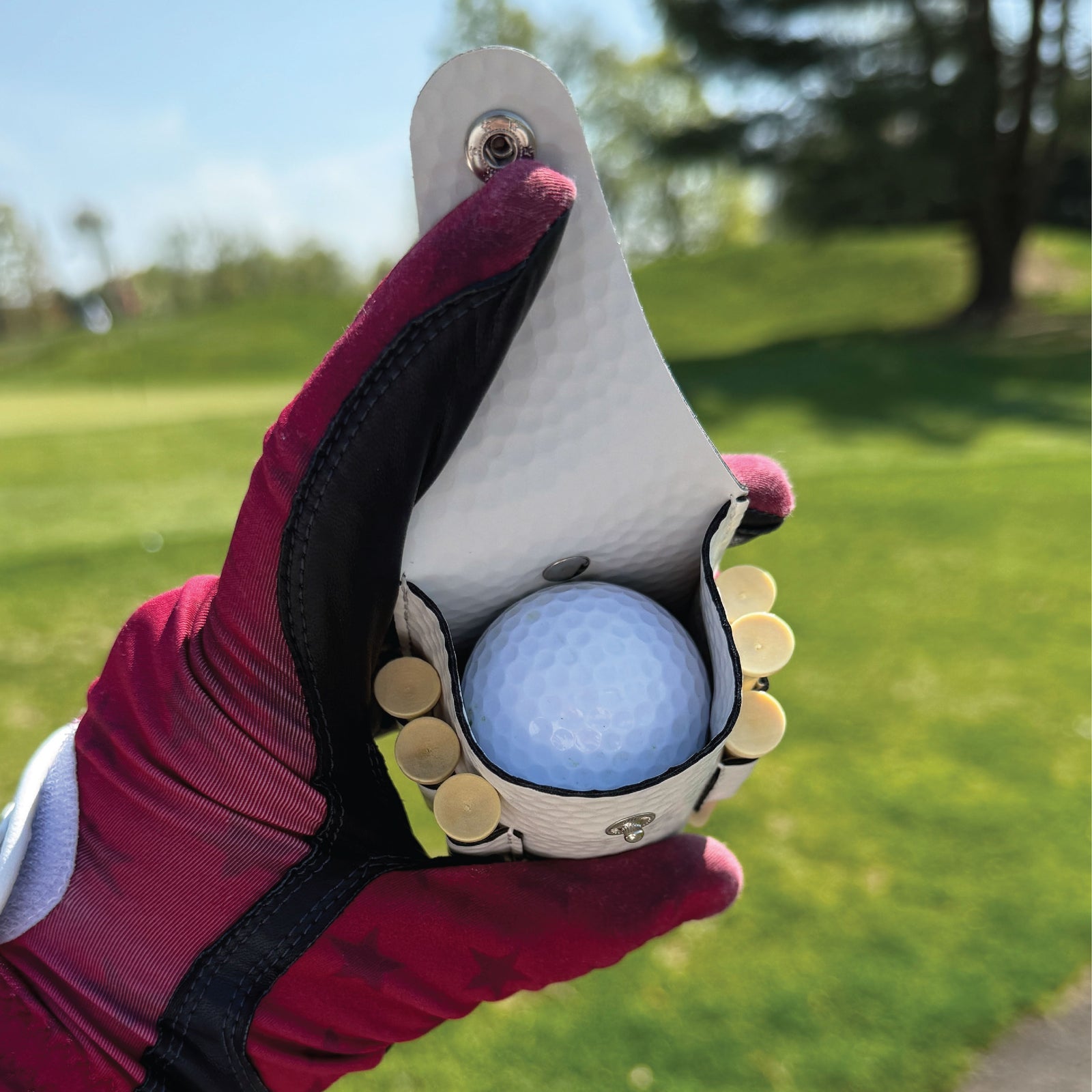 White leather golf ball pouch held in burgundy gloved hand, displaying blue golf ball and integrated ball markers on the course