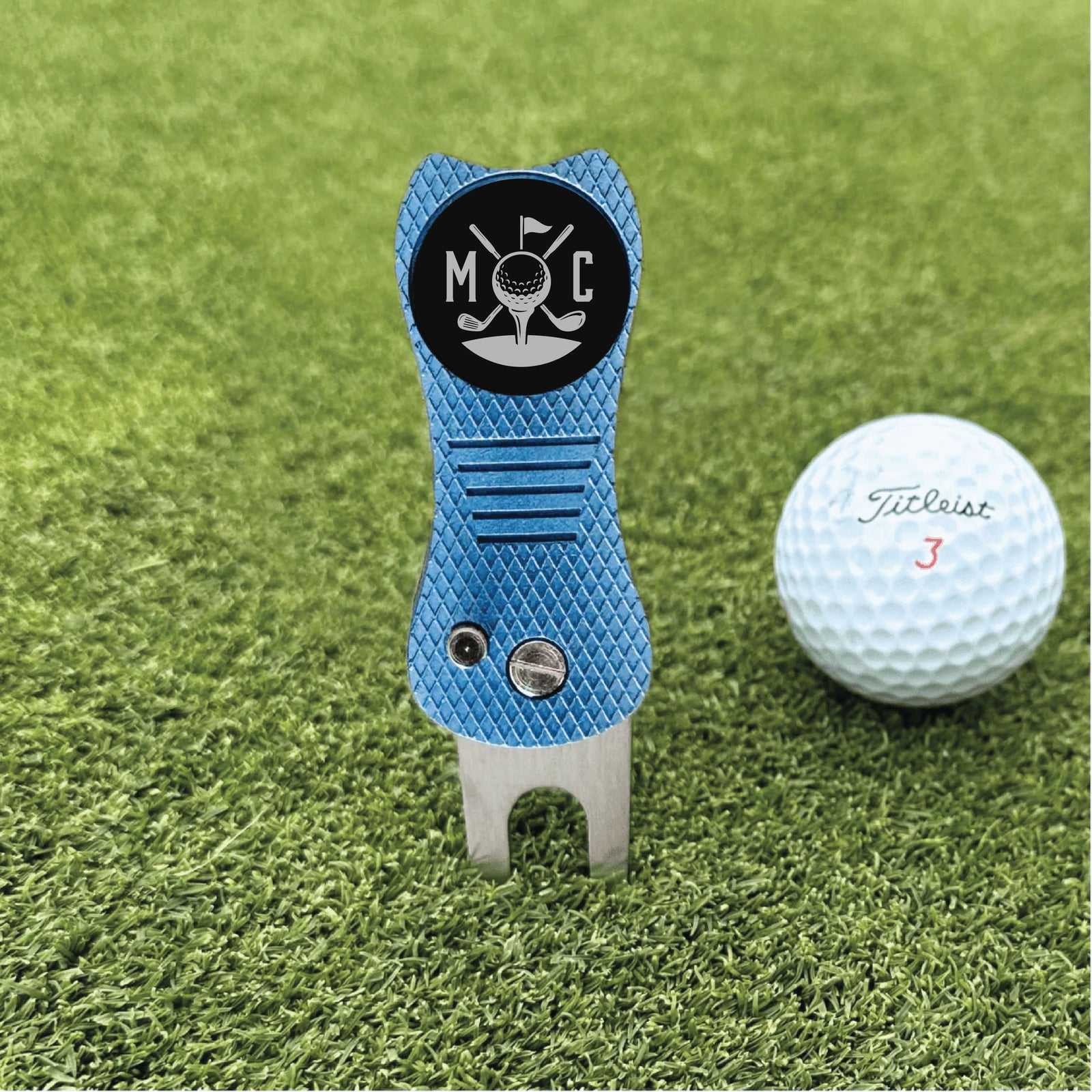 Blue divot tool with sample engraved initials "MC" and golf icon on black circular face, displayed on grass next to white golf ball