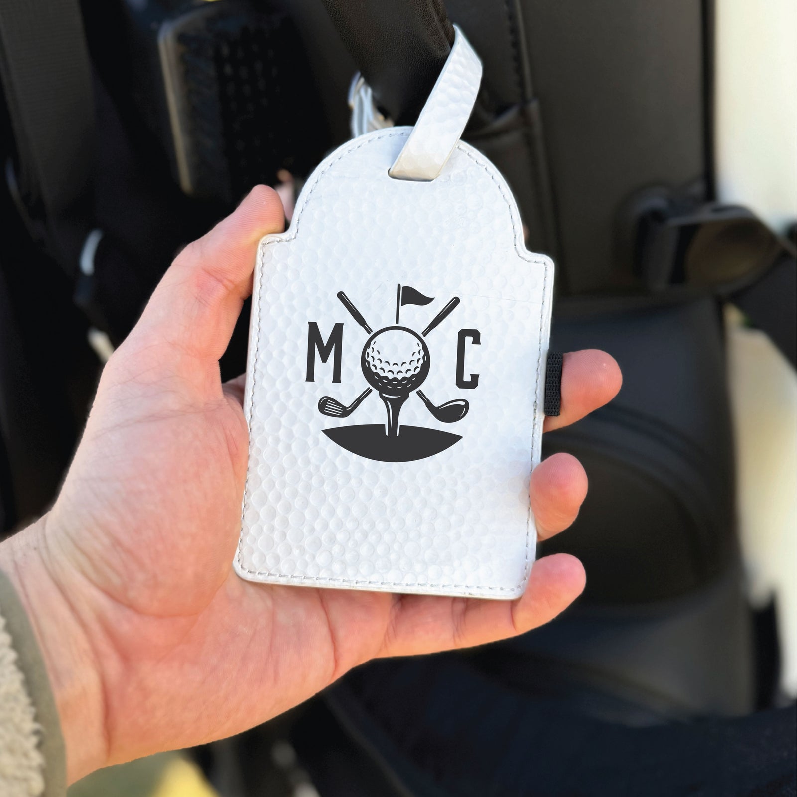 White quilted golf ball pouch with sample initials "MOC" and golf icon design featuring crossed clubs, ball on tee, and flag, held in hand with white ribbon attachment loop