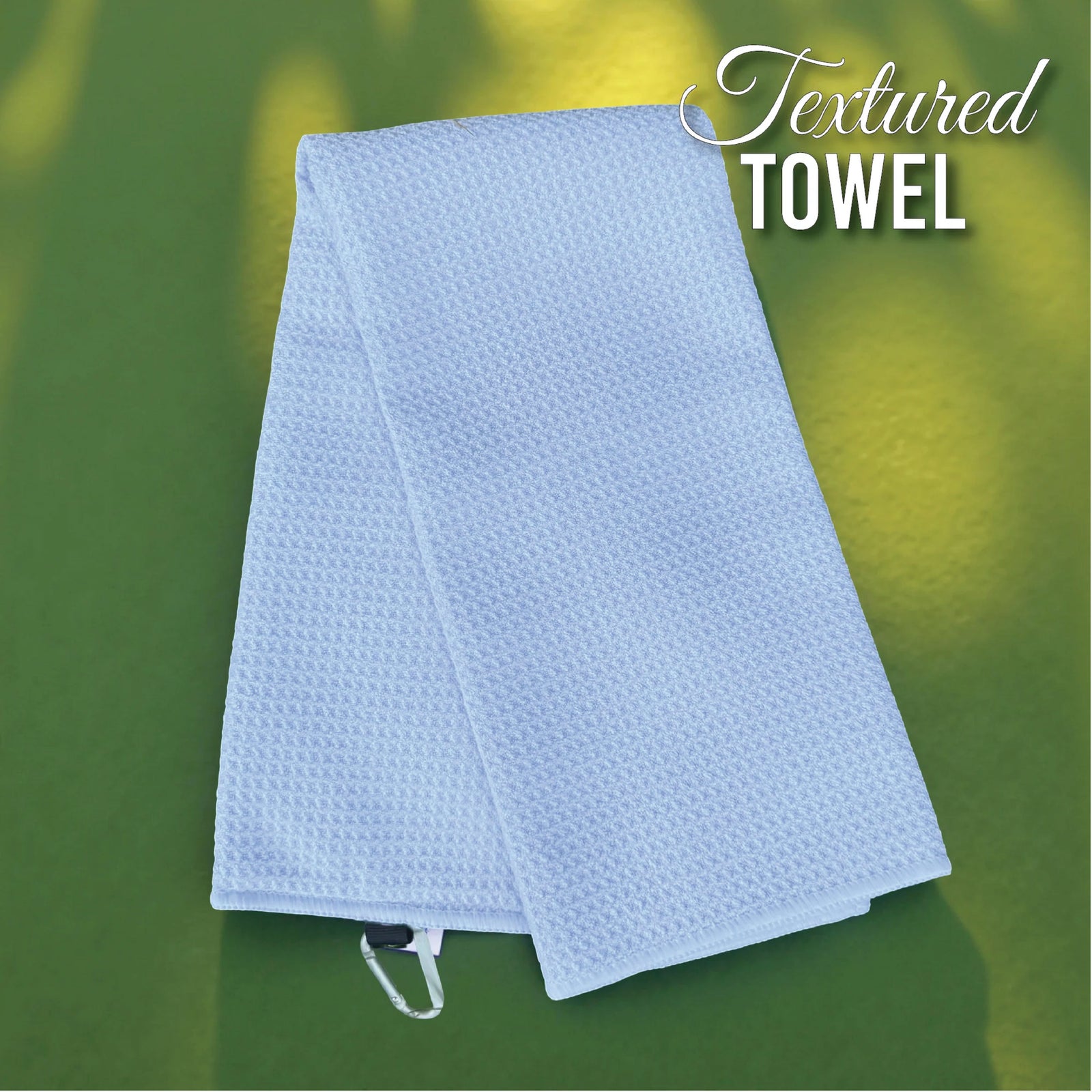 Light blue textured golf towel with waffle weave pattern and hanging loop, displayed against blurred green background
