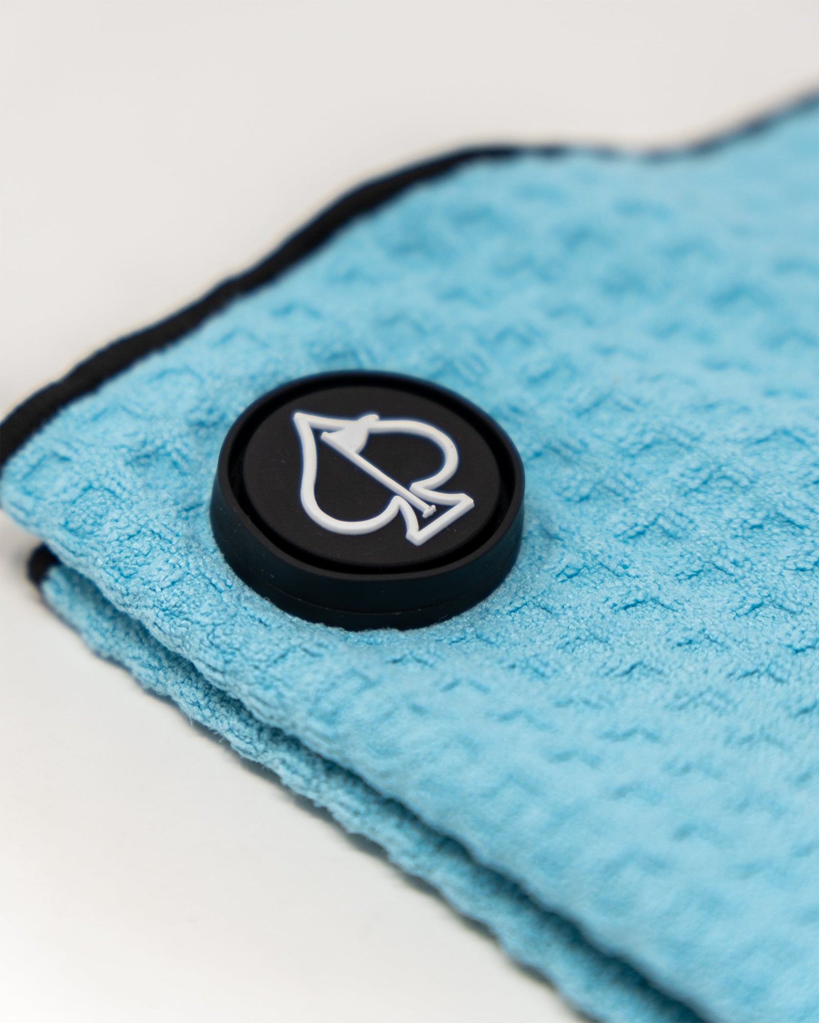 Light blue waffle-knit golf towel with black magnetic attachment featuring white spade icon emblem, close-up detail view