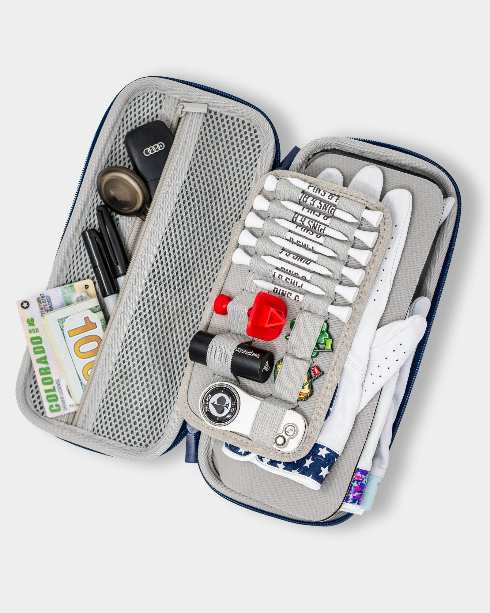 Black and navy glove caddie opened flat to display interior compartments holding white golf gloves, golf balls, tees with sample text, ball markers, divot tool, and other accessories