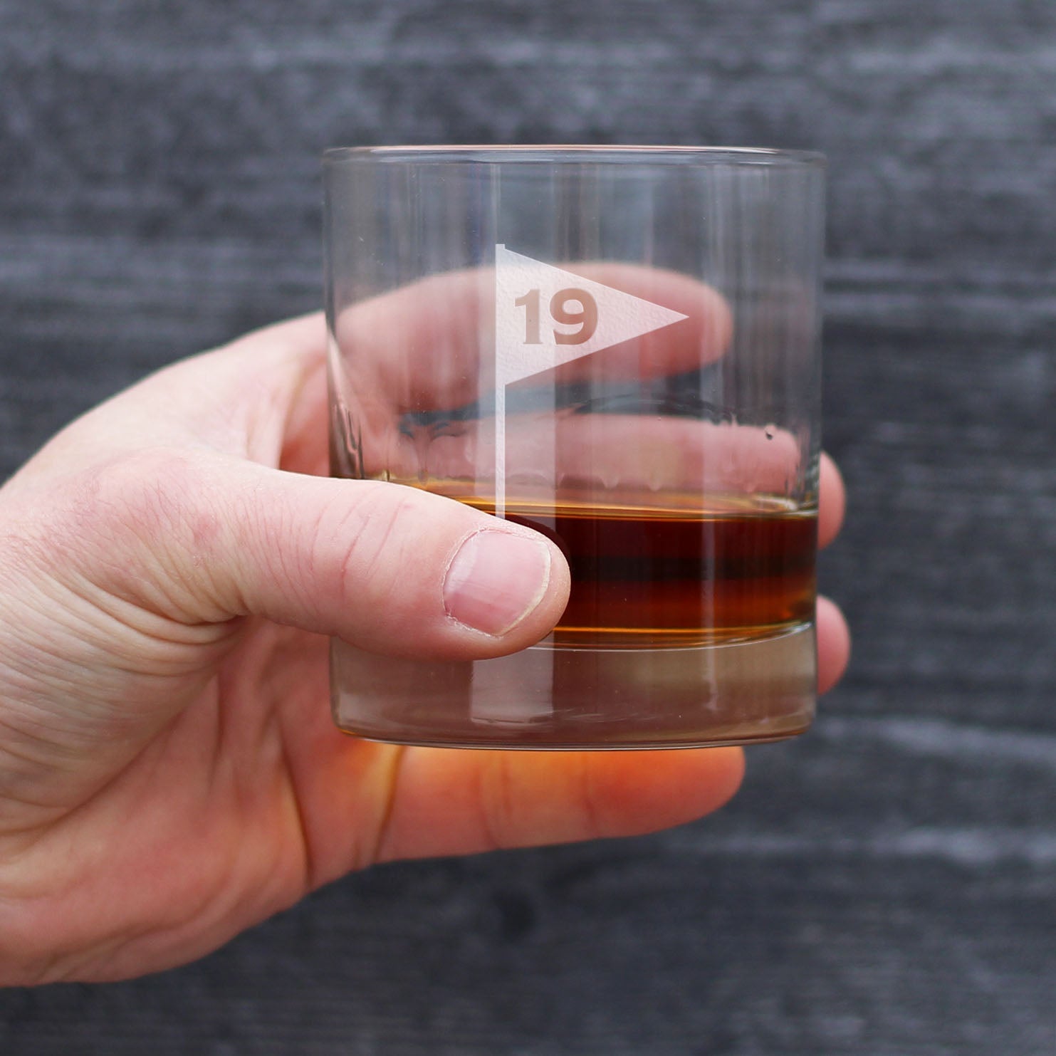 Hand holding rocks glass filled with amber liquid, engraved with golf flag icon and sample number 19
