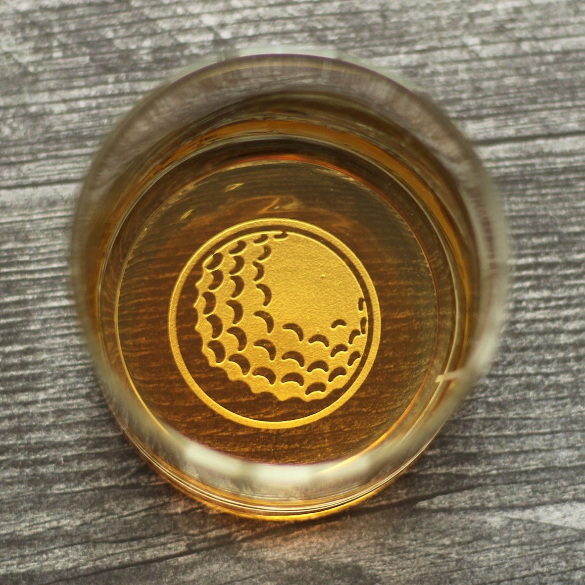 Gold-embossed golf ball design with dimple pattern centered on brown wooden base, viewed from above on gray wood surface.