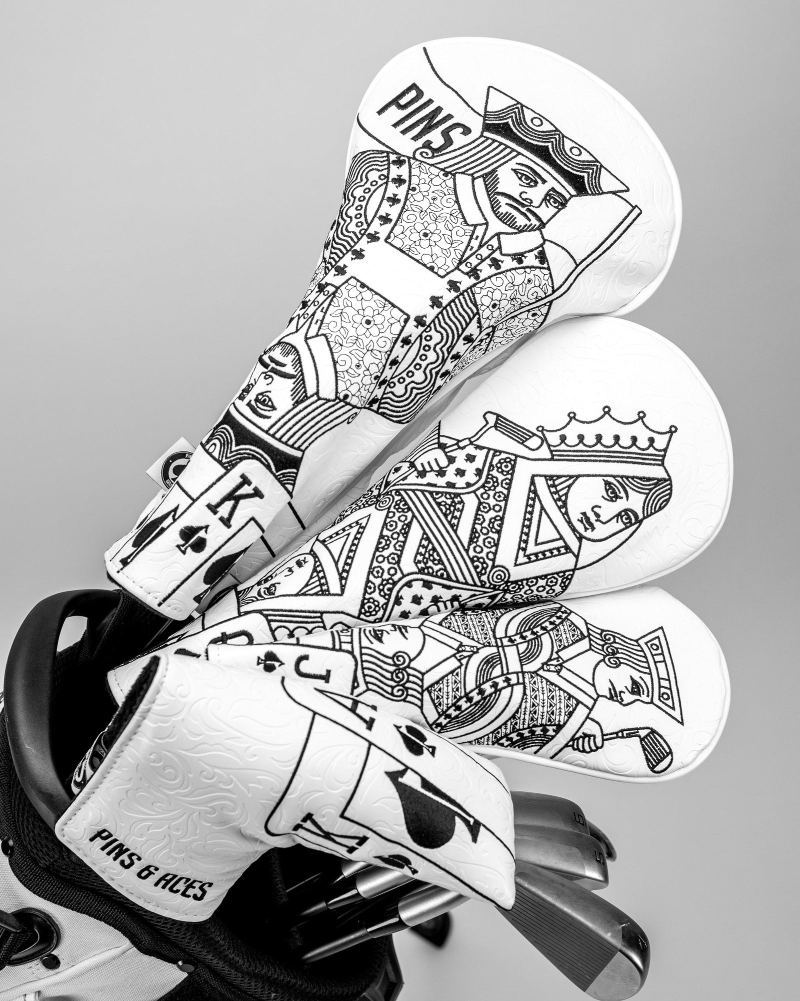 Stack of three white putter blade covers with black playing card designs, including Ace of Spades embroidery and "PINS & ACES" text on the base cover