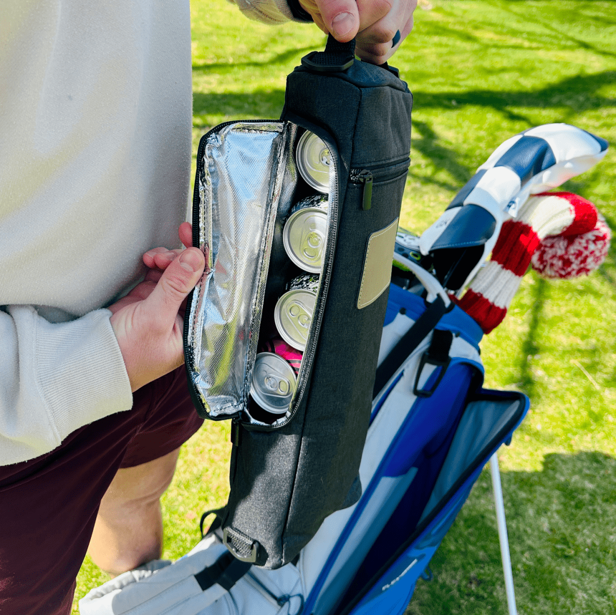 Black insulated cooler bag opened to show interior compartment holding four beverage cans, displayed on golf course with clubs and head covers in background
