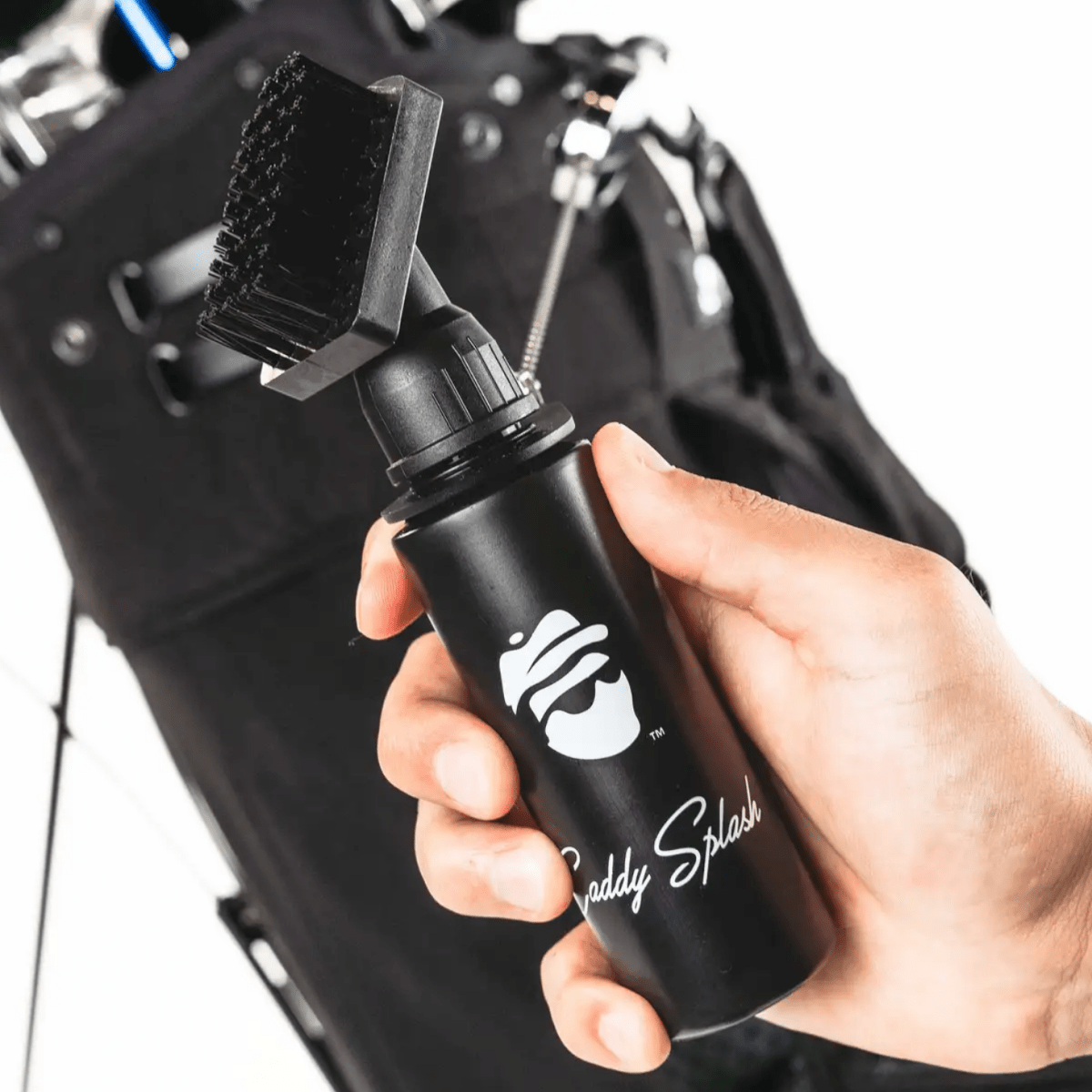 Black golf towel spray bottle with white logo and "Caddy Splash" text, held in hand over golf bag in background