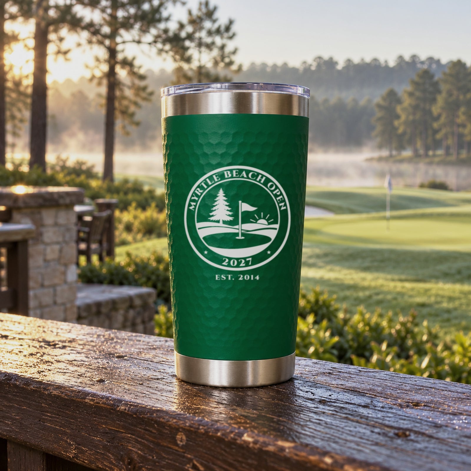 Green golf ball tumbler engraved with a golf club seal reading Myrtle Beach Open 2027, a personalized golf trip gift on a clubhouse rail