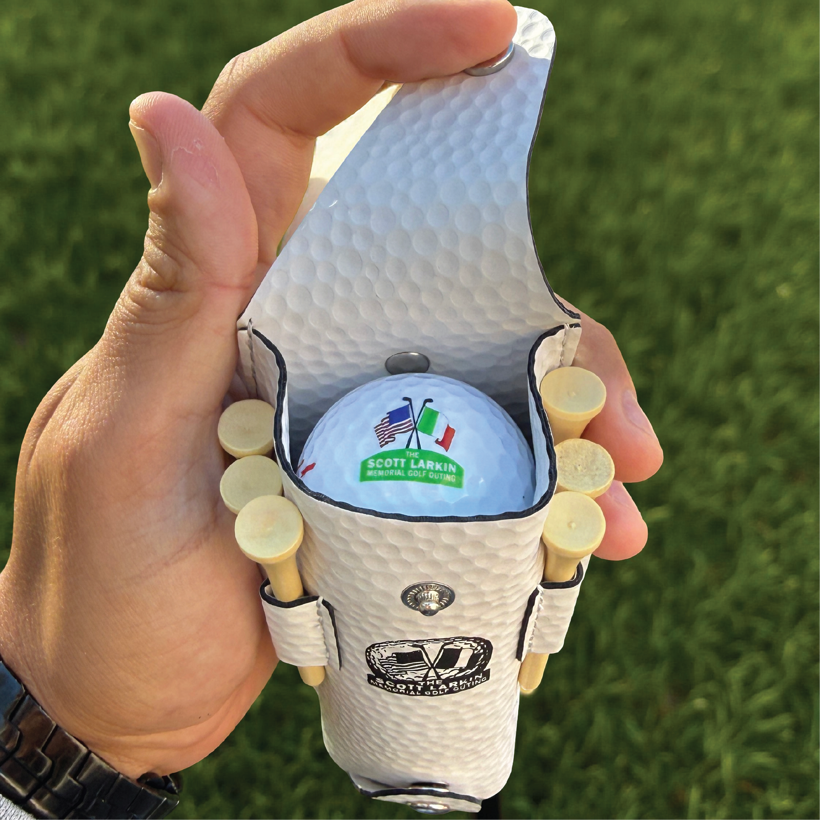 White golf ball-textured pouch held open in hand, displaying sample-printed golf ball with "SCOTT LARKIN" text and flag logos, with wooden tees visible in side pockets