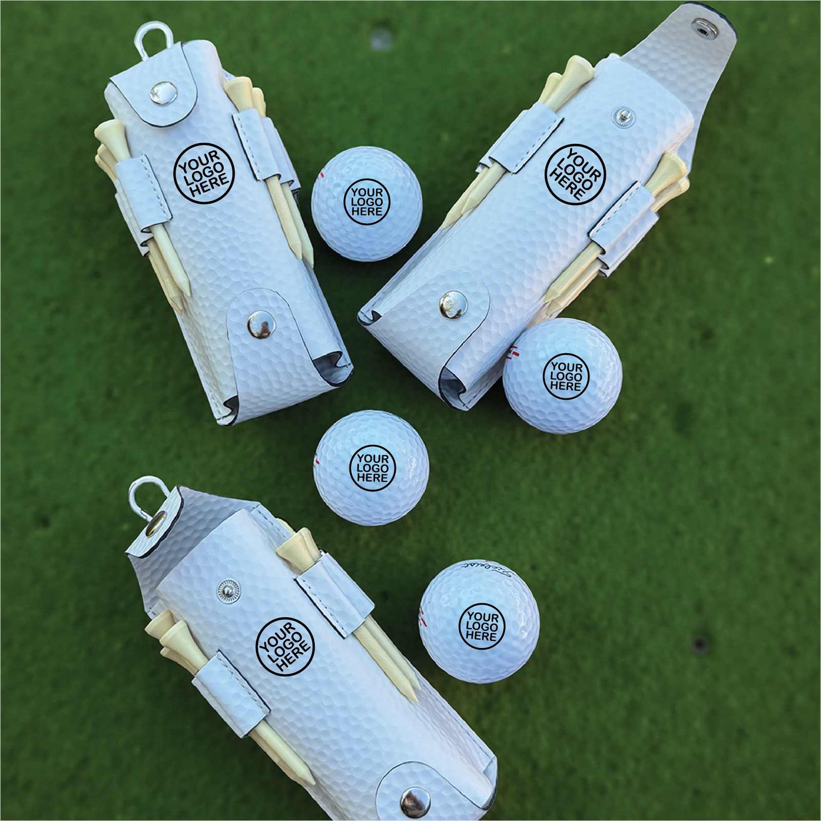 Flat-lay overhead view of light blue golf accessory sets on grass showing white pouches with sample logo imprint, included golf balls with sample logo placement, and wooden tees