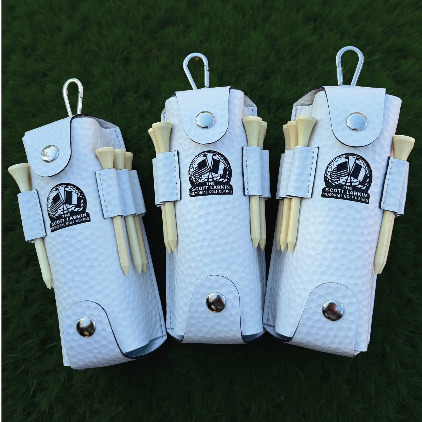 Three white textured pouches with carabiner clips, each holding wooden tees and displaying the Scott Larkin Memorial Golf Outing logo, arranged on a grass background.