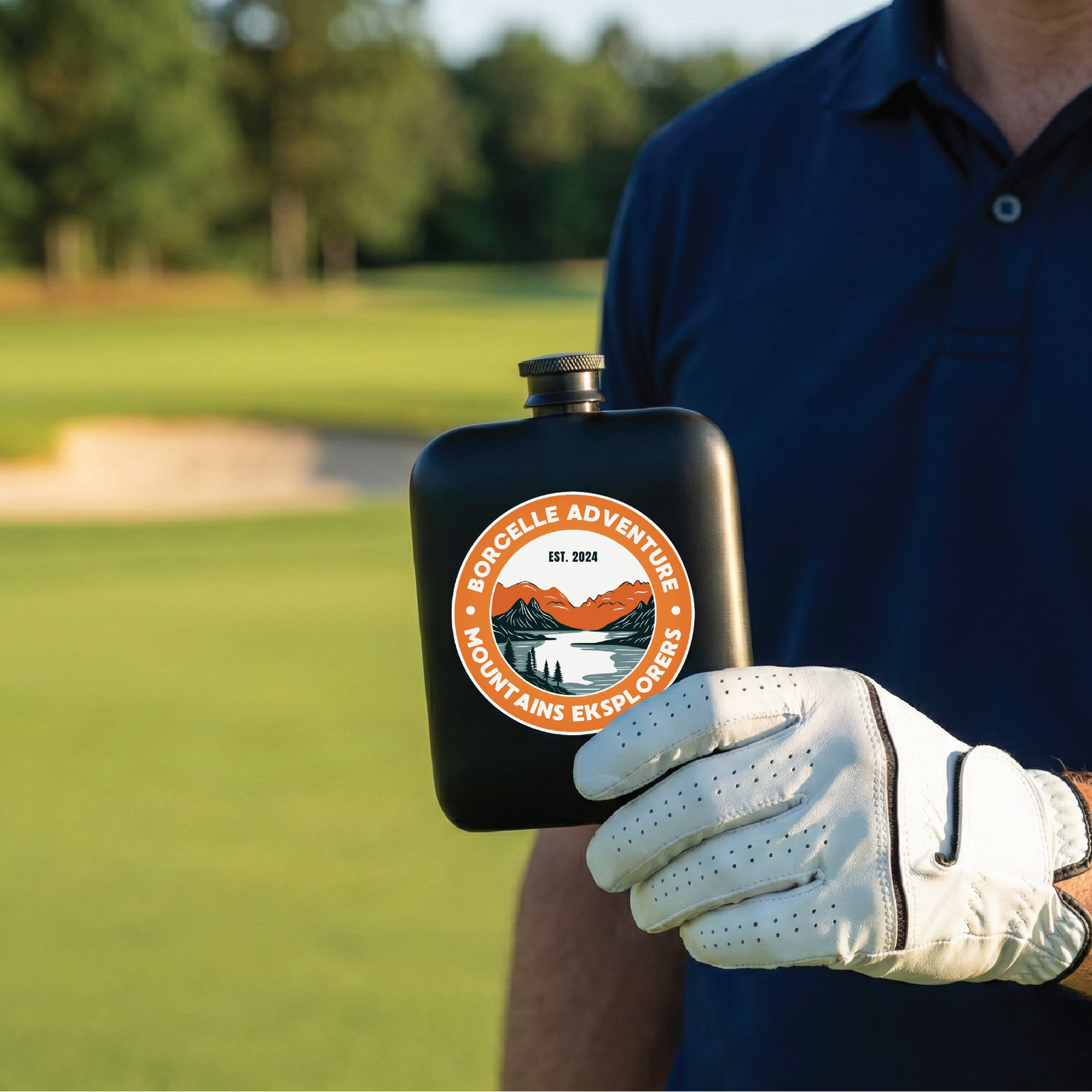 Black stainless steel flask with orange and white circular logo reading "Borcelle Adventure Mountains Explorers Est. 2024" held in golfer's hand wearing white glove on golf course