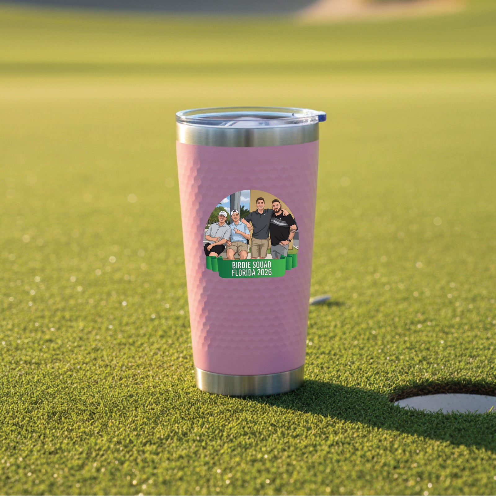 Pink insulated tumbler with stainless steel band displayed on golf course green, featuring personalized photo graphic with sample names "Birdie Squad Florida 2026