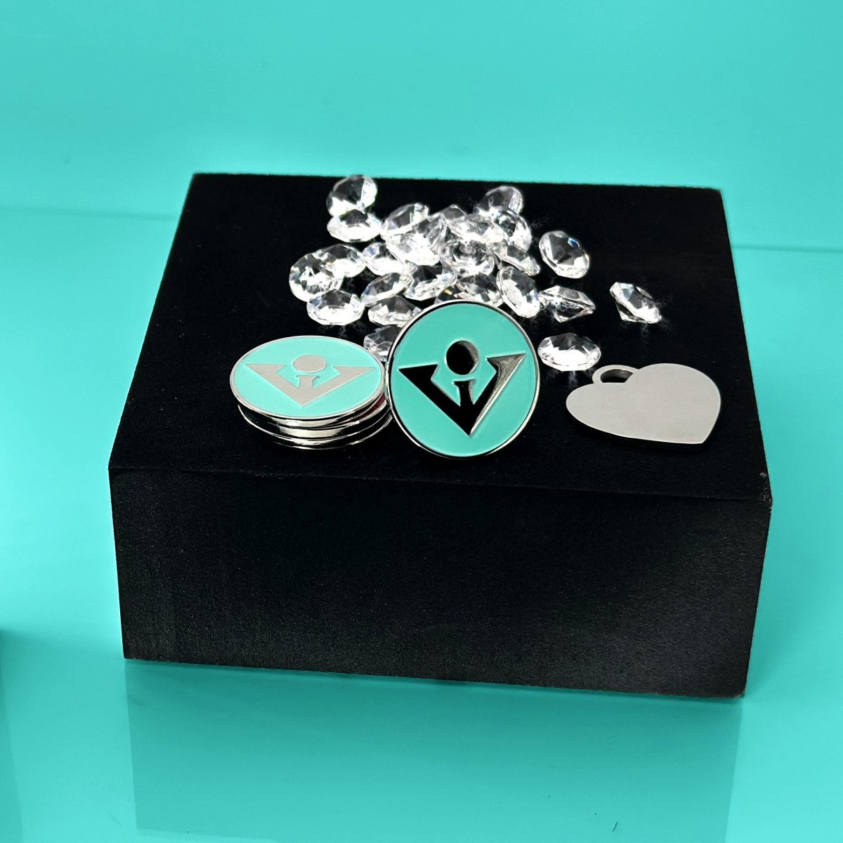 Collection of ball markers displayed on black box, including turquoise round marker with logo, stacked square markers, heart-shaped marker, and scattered crystal decorative elements against turquoise background