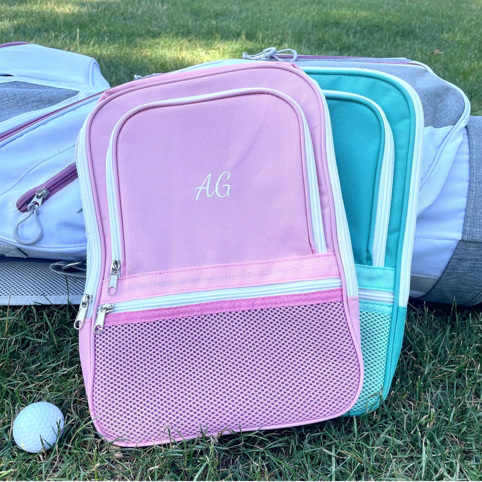 Pink golf shoe bag with sample embroidered initials "AG" on front panel, displayed alongside turquoise version on grass with golf balls nearby