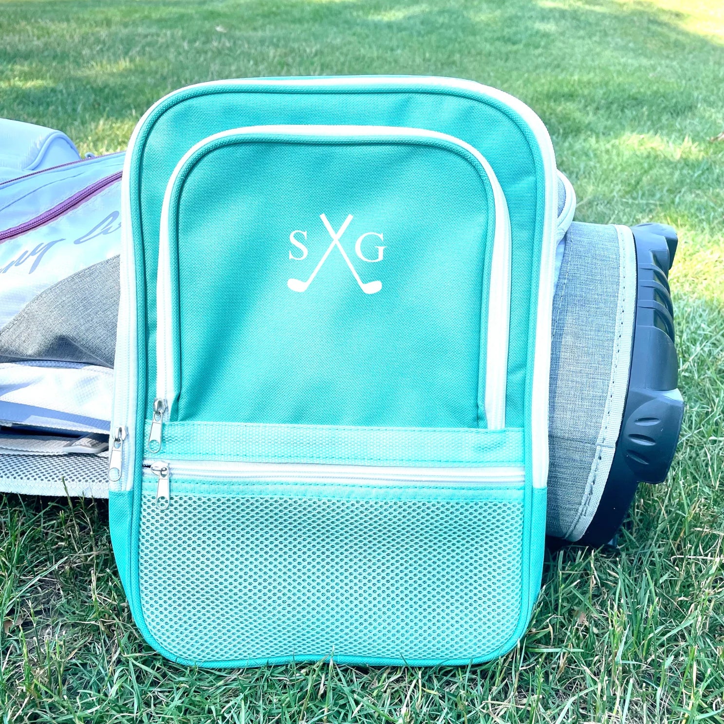 Turquoise shoe bag with white trim and mesh pocket, embroidered with sample initials "sXG" on the main compartment, displayed on grass beside golf equipment.