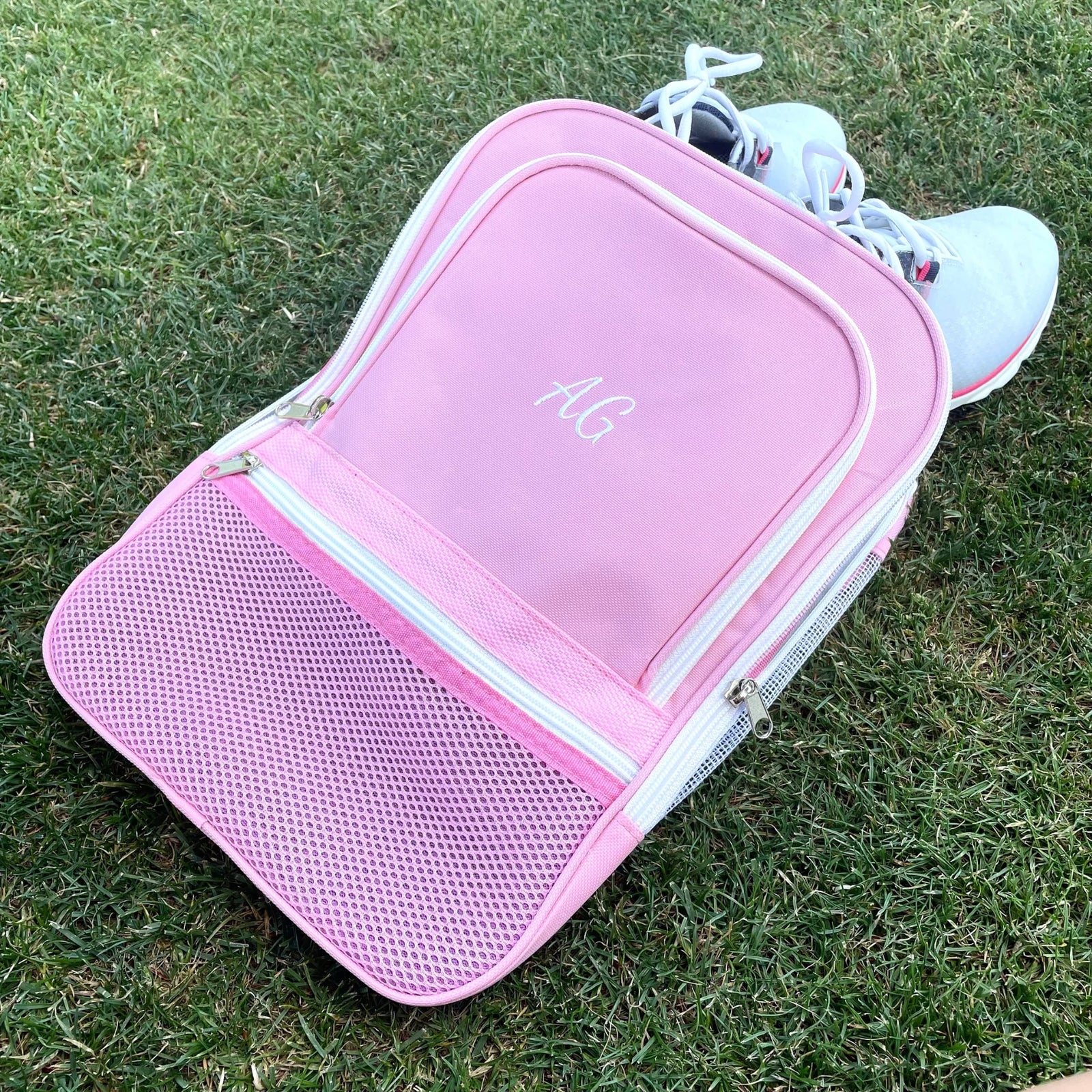 Pink golf shoe bag with embroidered sample initials "ALC" on main compartment, displayed on grass with white golf shoes nearby