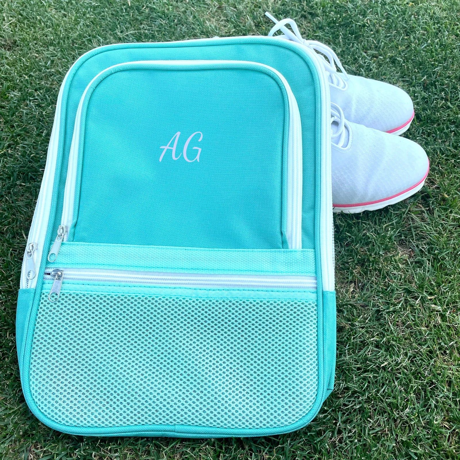 Turquoise canvas shoe bag with white trim and mesh pocket, embroidered with sample initials "AG", displayed on grass next to white golf shoes with pink accents