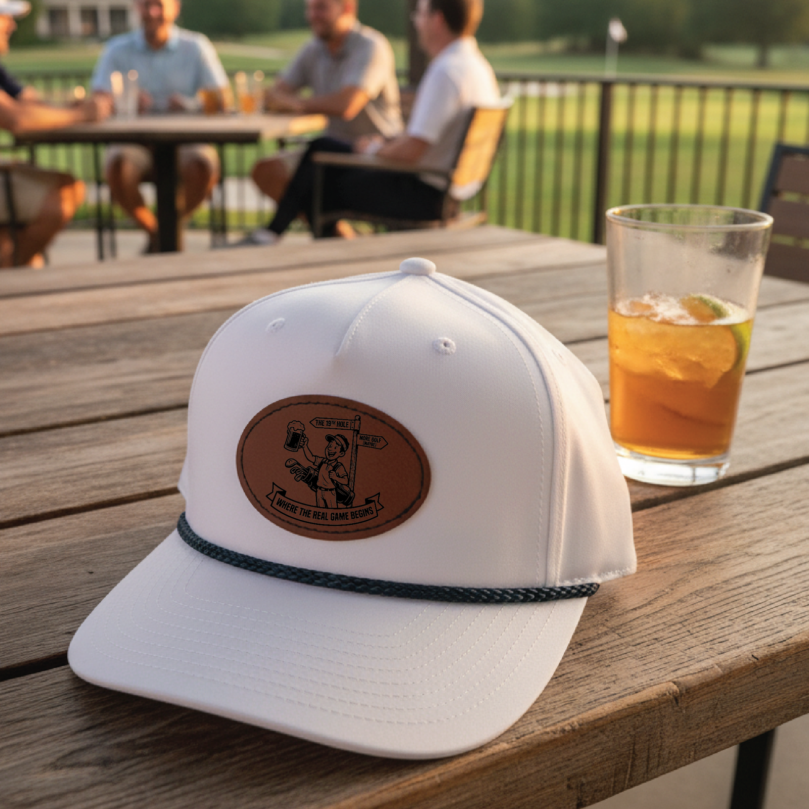 White golf hat with brown leather patch featuring engraved illustration and text, displayed on deck table with beverage nearby and golfers in background.