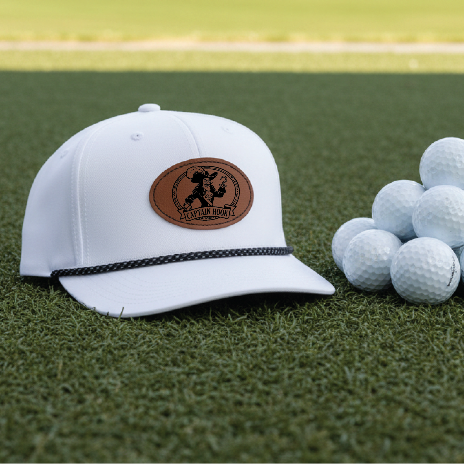 White golf polo hat with brown leather patch featuring "Captain Hook" emblem and black rope trim, displayed on putting green next to golf balls