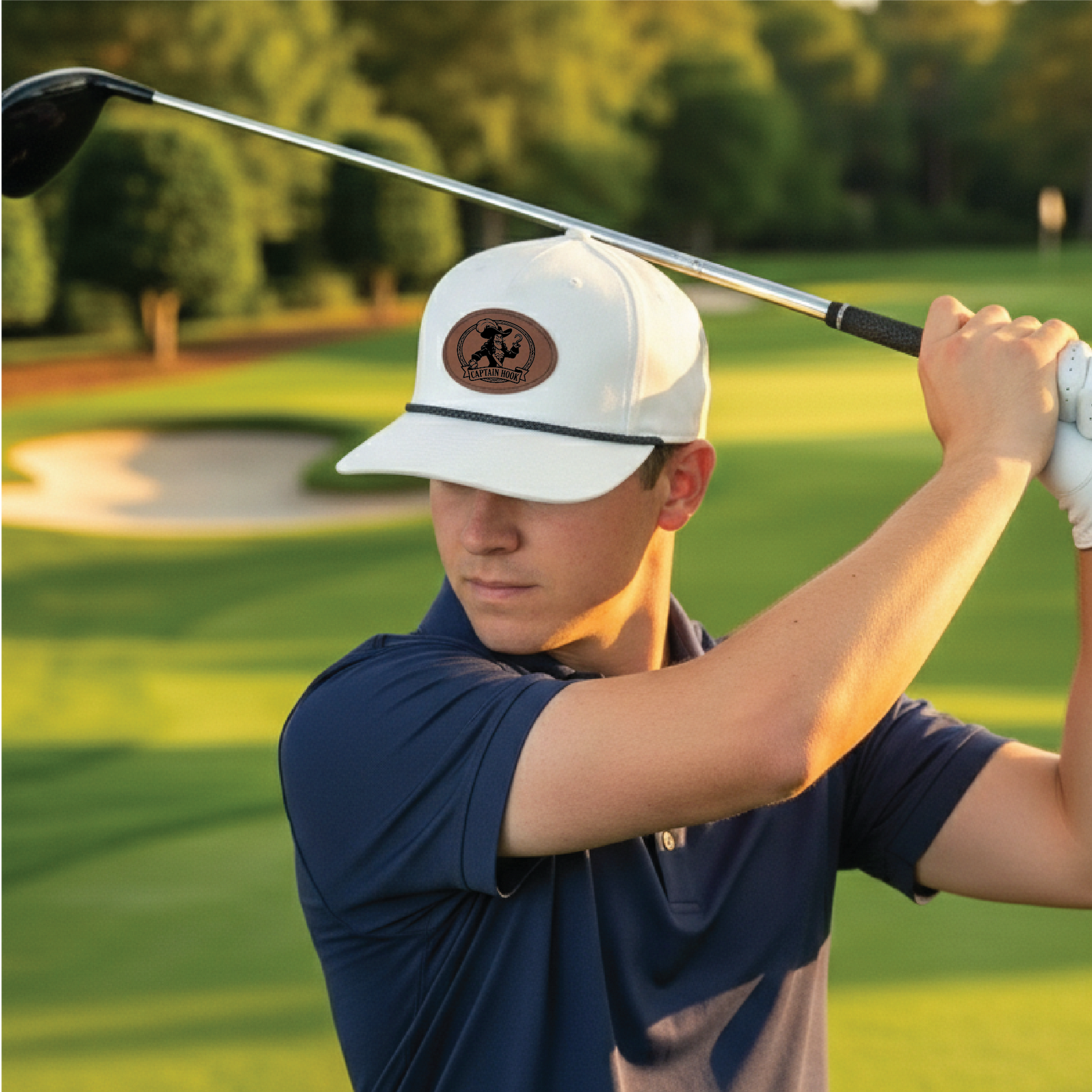 White golf cap with brown leather patch featuring sample text "Captain Hook" worn on golf course during swing, navy polo visible