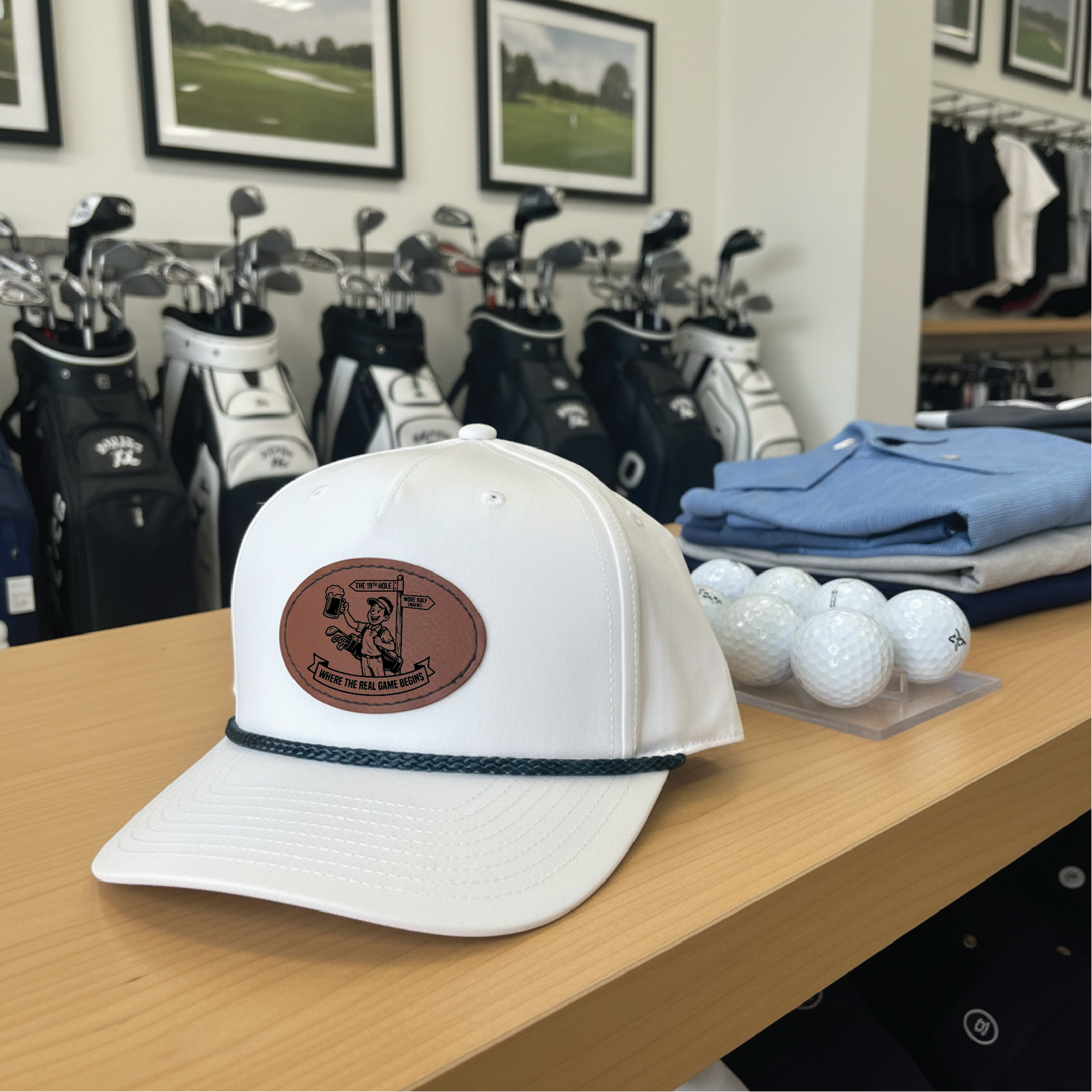 White golf hat with brown leather patch featuring golfer illustration and text, displayed on wooden table in pro shop setting with golf bags and accessories visible in background