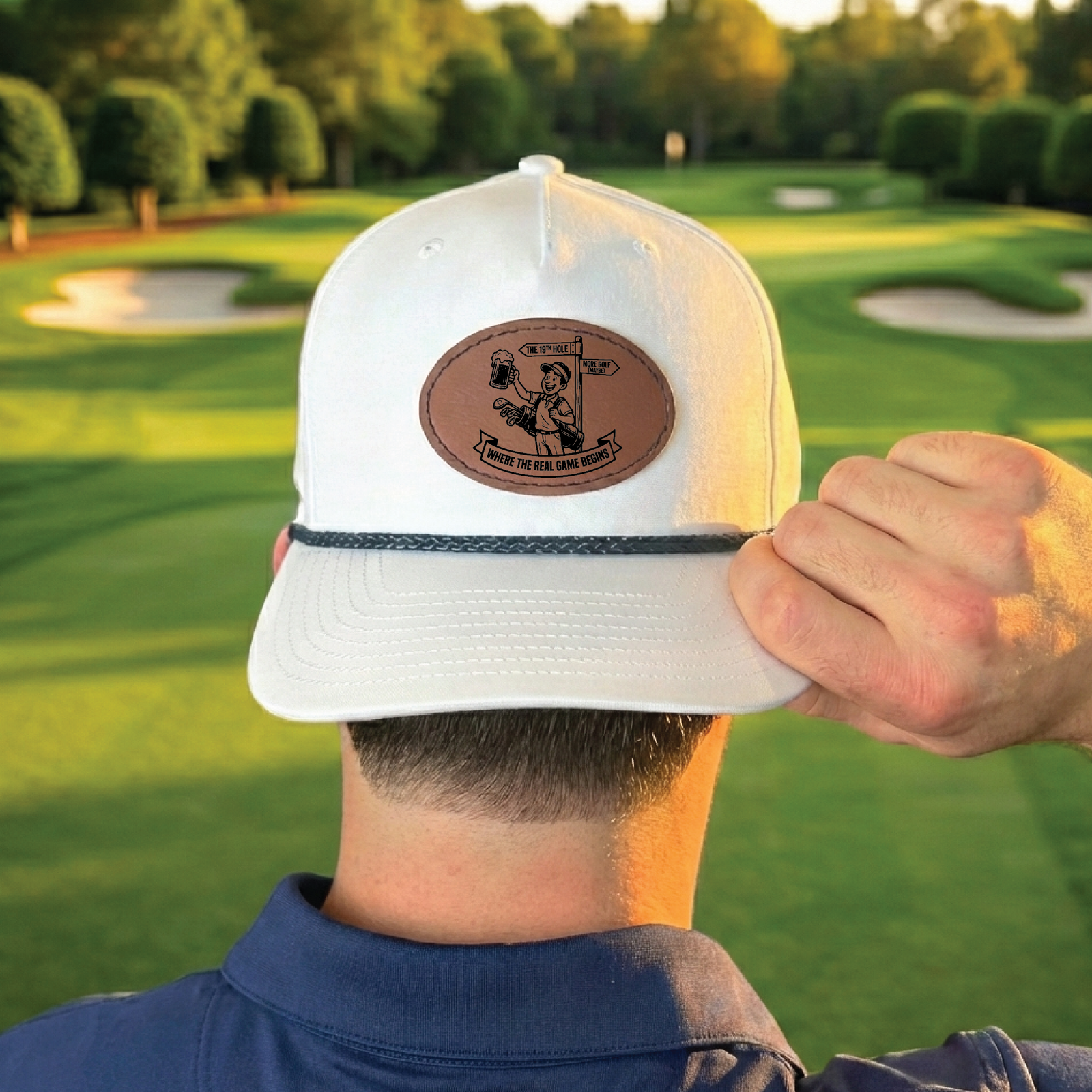 White baseball cap with brown leather patch featuring engraved golf-themed illustration and text, worn on golfer's head with golf course in background.