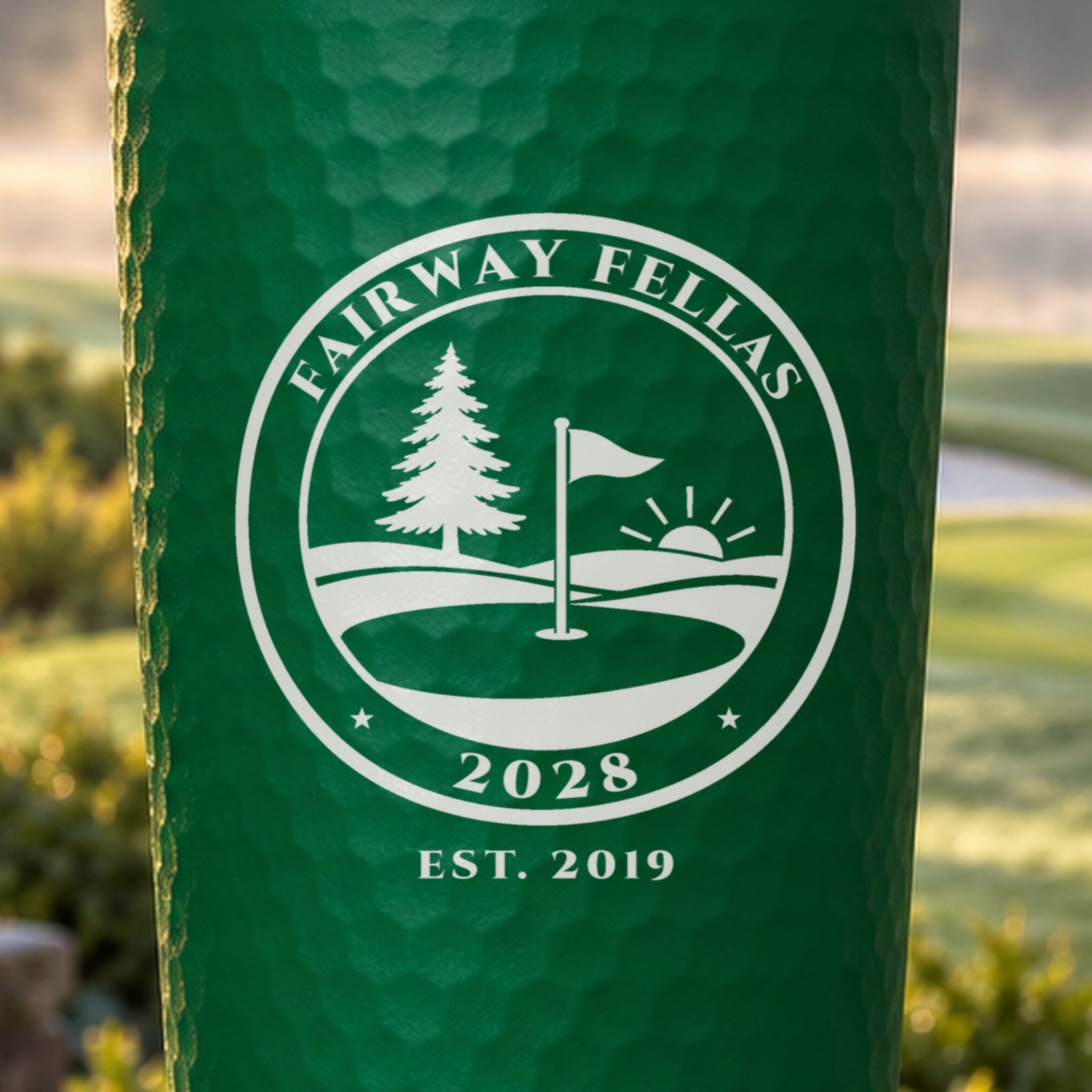 Close-up of the laser engraved golf club seal on a green dimpled tumbler reading Fairway Fellas 2028 with a pine, flagstick and sunrise