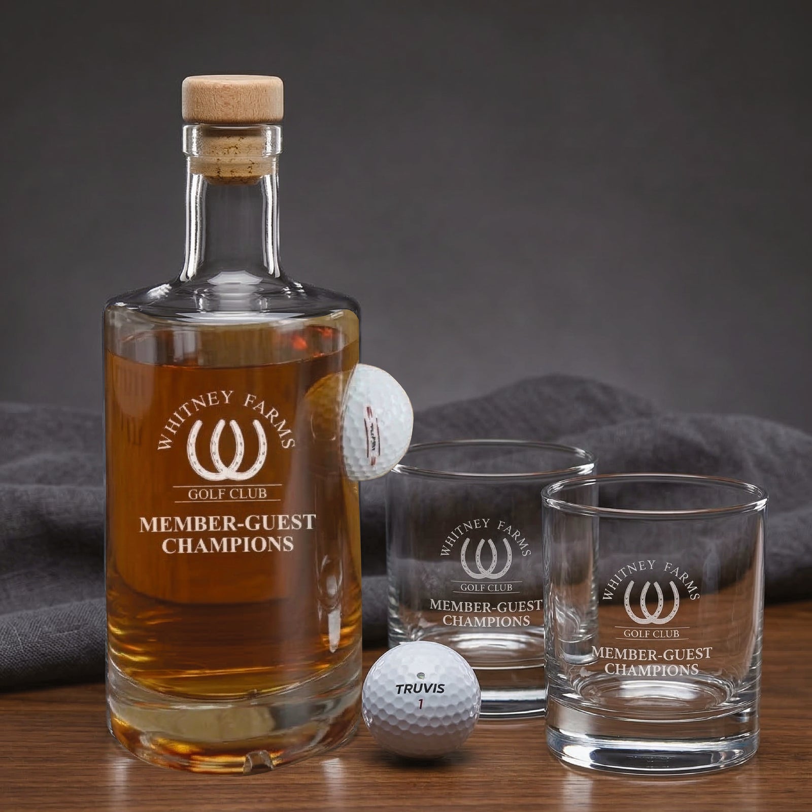Golf decanter set with cork-topped bottle filled with amber whiskey, two matching glasses, and two golf balls engraved with "Whitney Farms Golf Club Member-Guest Champions" logo