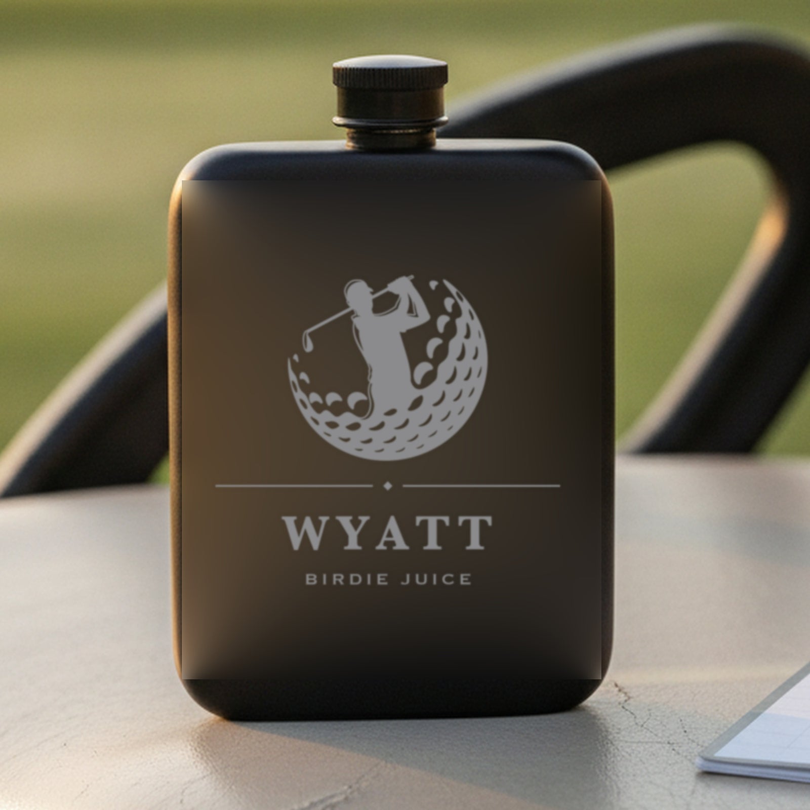 Engraved golf flask reading Birdie Juice standing on a golf cart seat beside a scorecard with a sunlit fairway behind it.