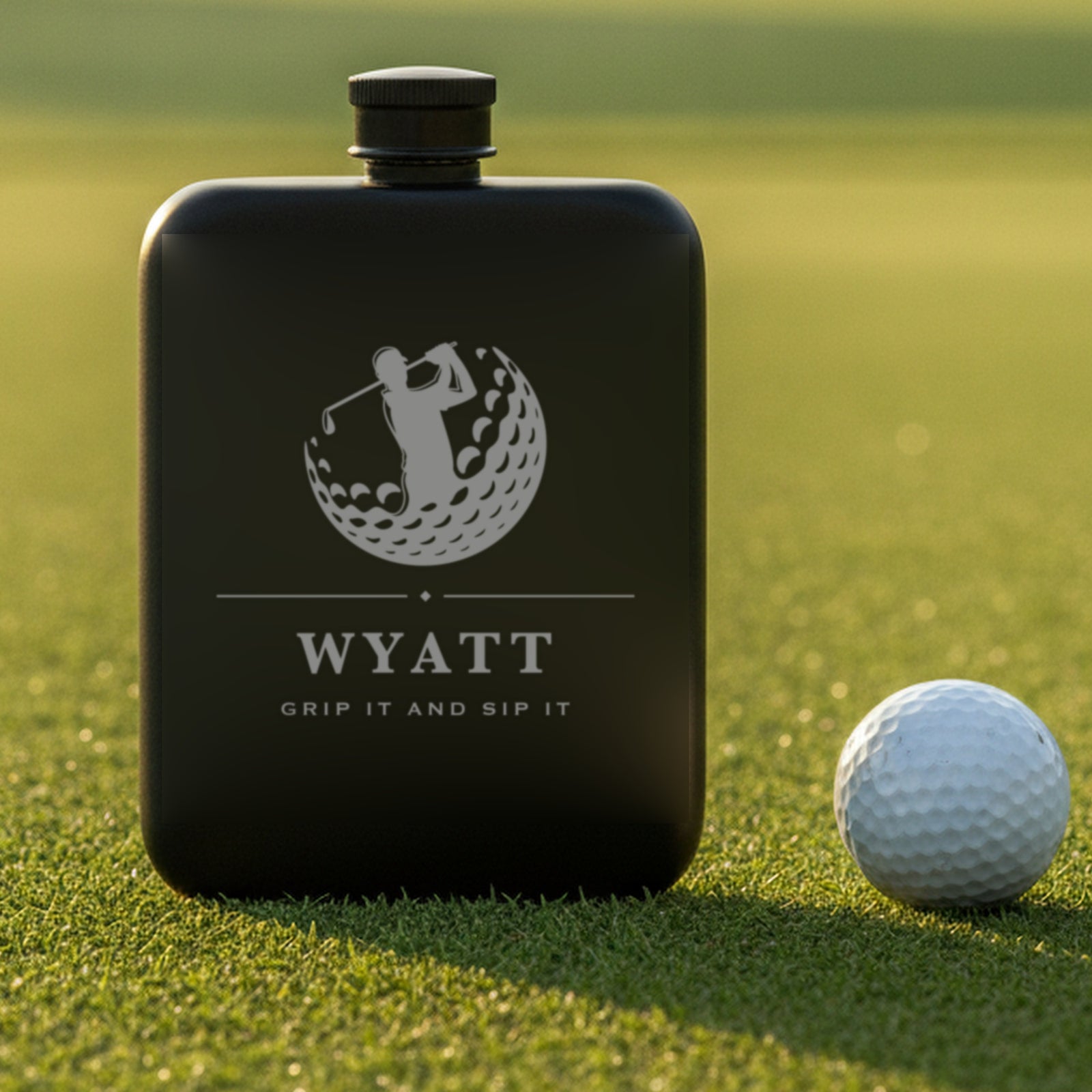 Personalized golf flask reading Grip It And Sip It standing on putting green turf beside a white golf ball at sunrise.