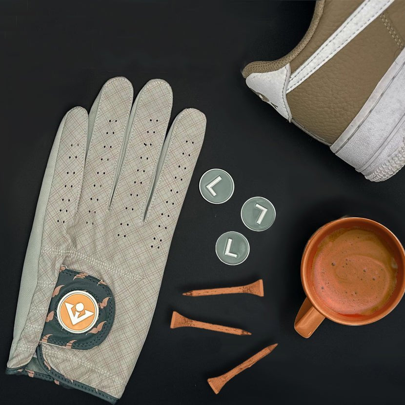 Cream Cabretta leather golf glove with mustache pattern displayed flat, showing perforated palm and reinforced patch, styled with wooden tees, ball markers, and golf accessories on dark background.