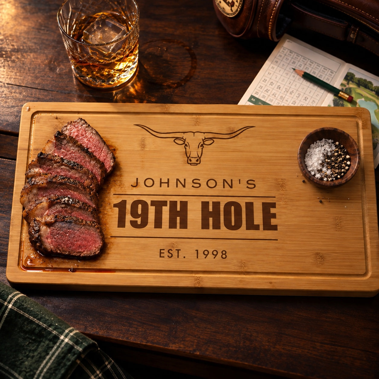 Bamboo cutting board engraved Johnson's 19th Hole Est. 1998 with a longhorn, sliced steak, whiskey and a golf scorecard on a bar top, a personalized golfer gift